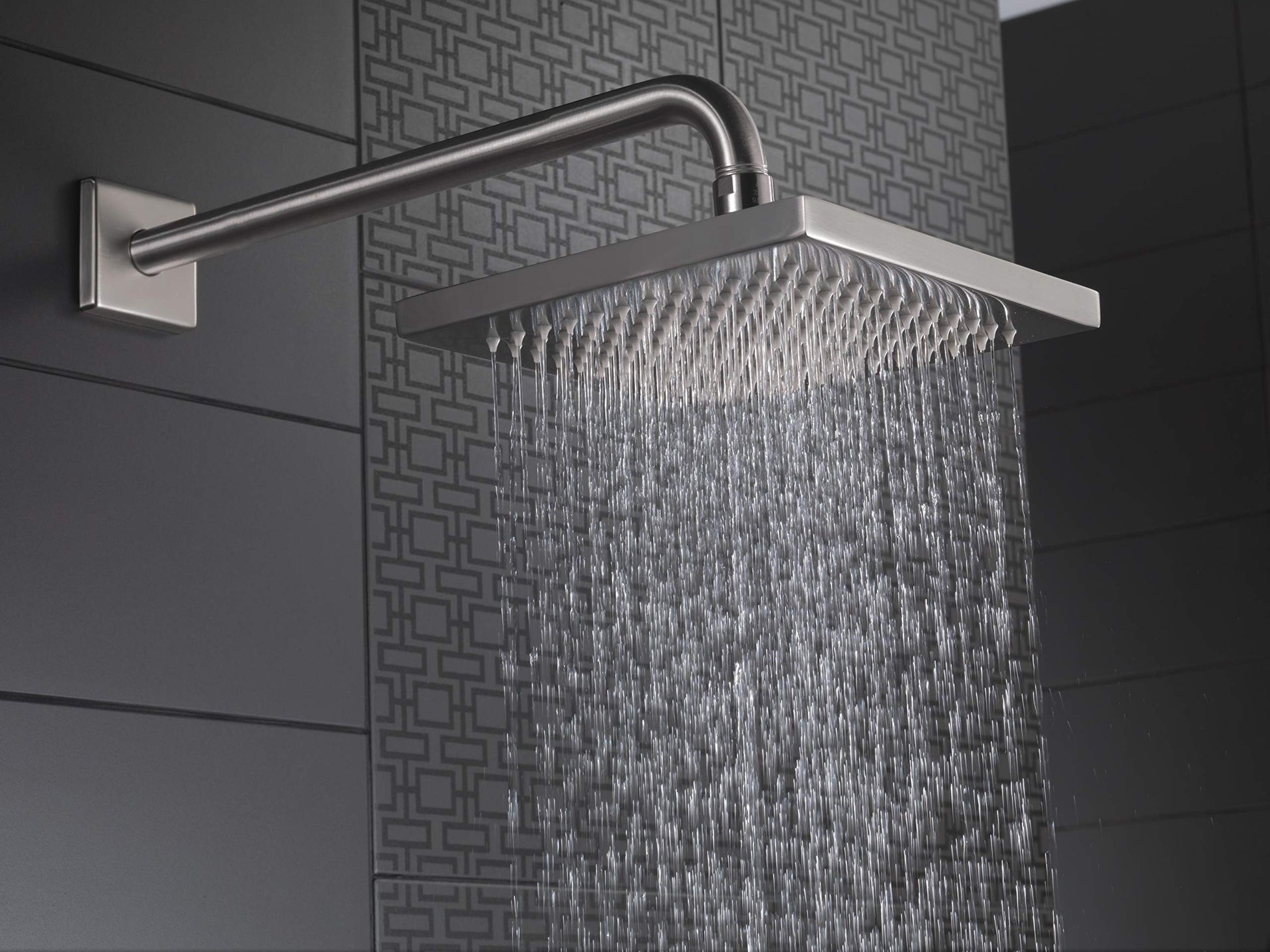 Stainless steel raincan shower head mounted on wall with curved arm, dispensing water in rain-like pattern.