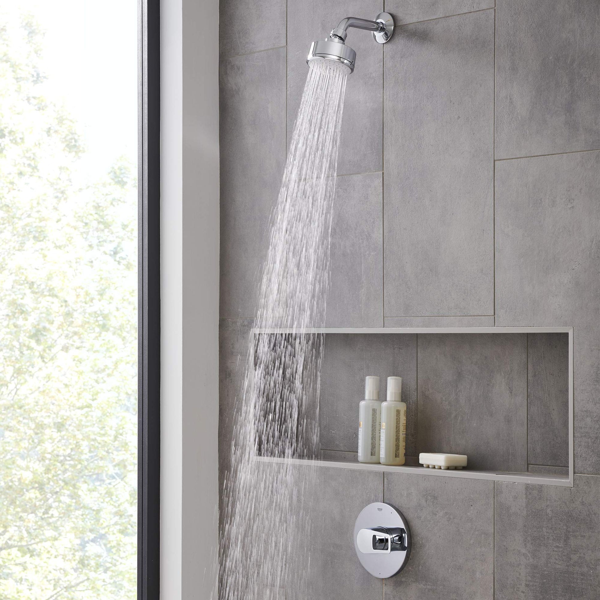 Chrome pressure balance valve trim with round handle and overhead rainfall showerhead mounted on gray tile wall.