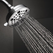Chrome shower head with multiple spray holes and rainfall stream pattern against gray tile wall.