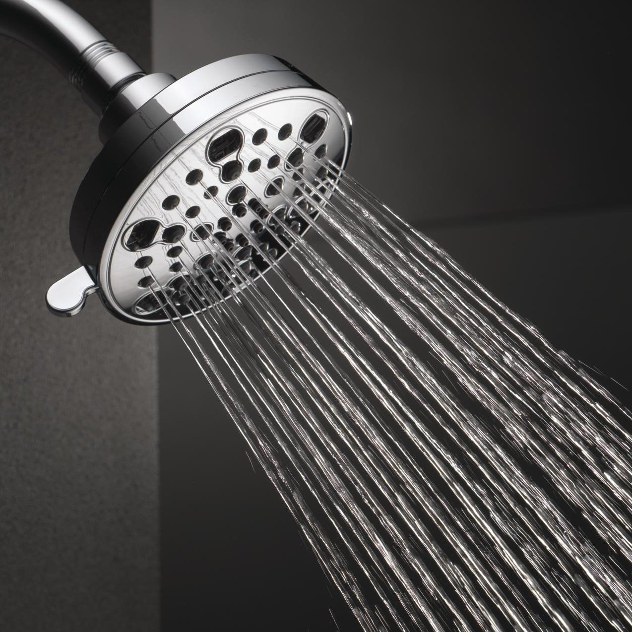 Chrome shower head with multiple spray holes and rainfall stream pattern against gray tile wall.
