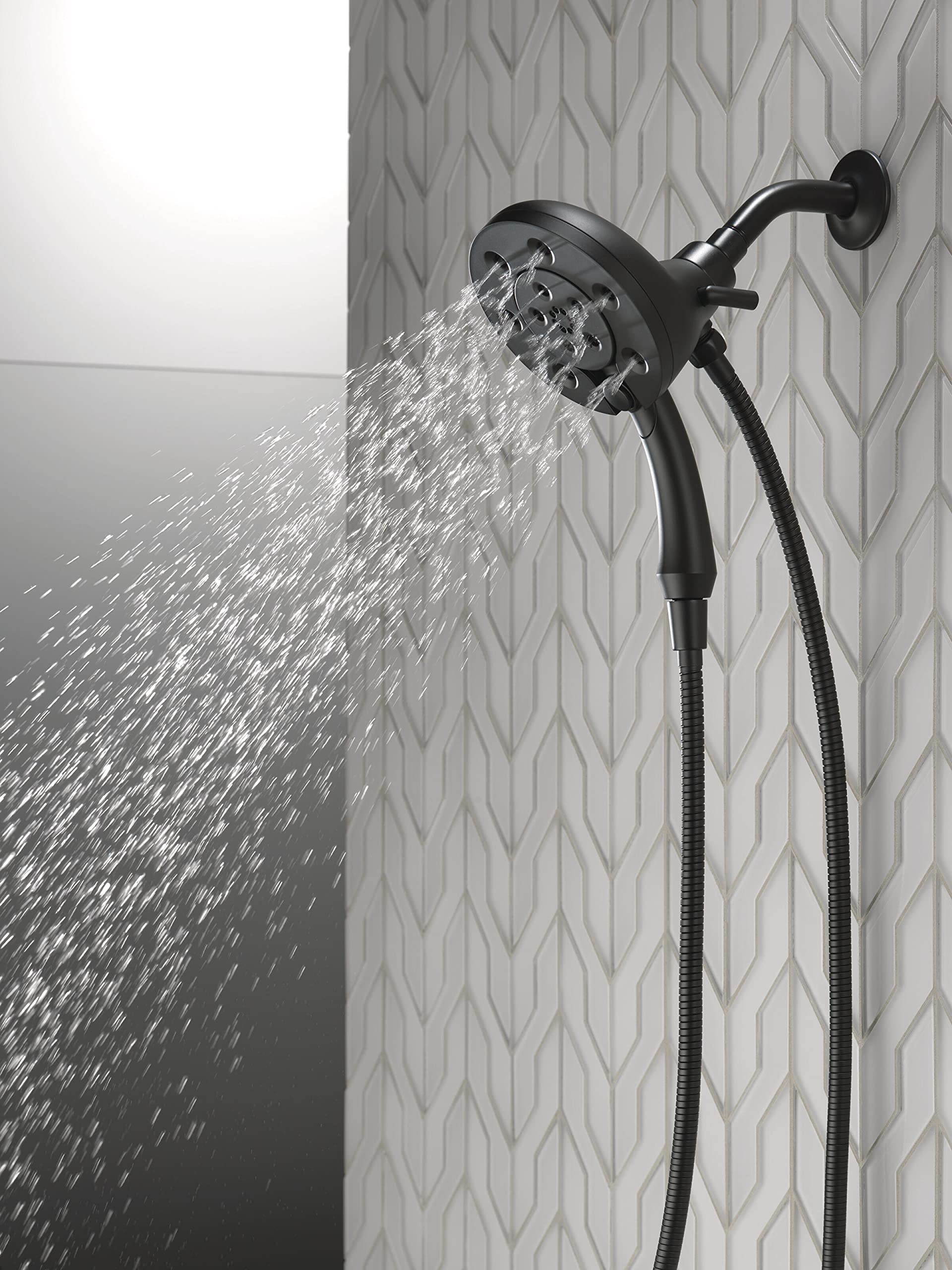 Matte black dual showerhead with water streaming, mounted on geometric patterned tile wall with flexible hose attached.