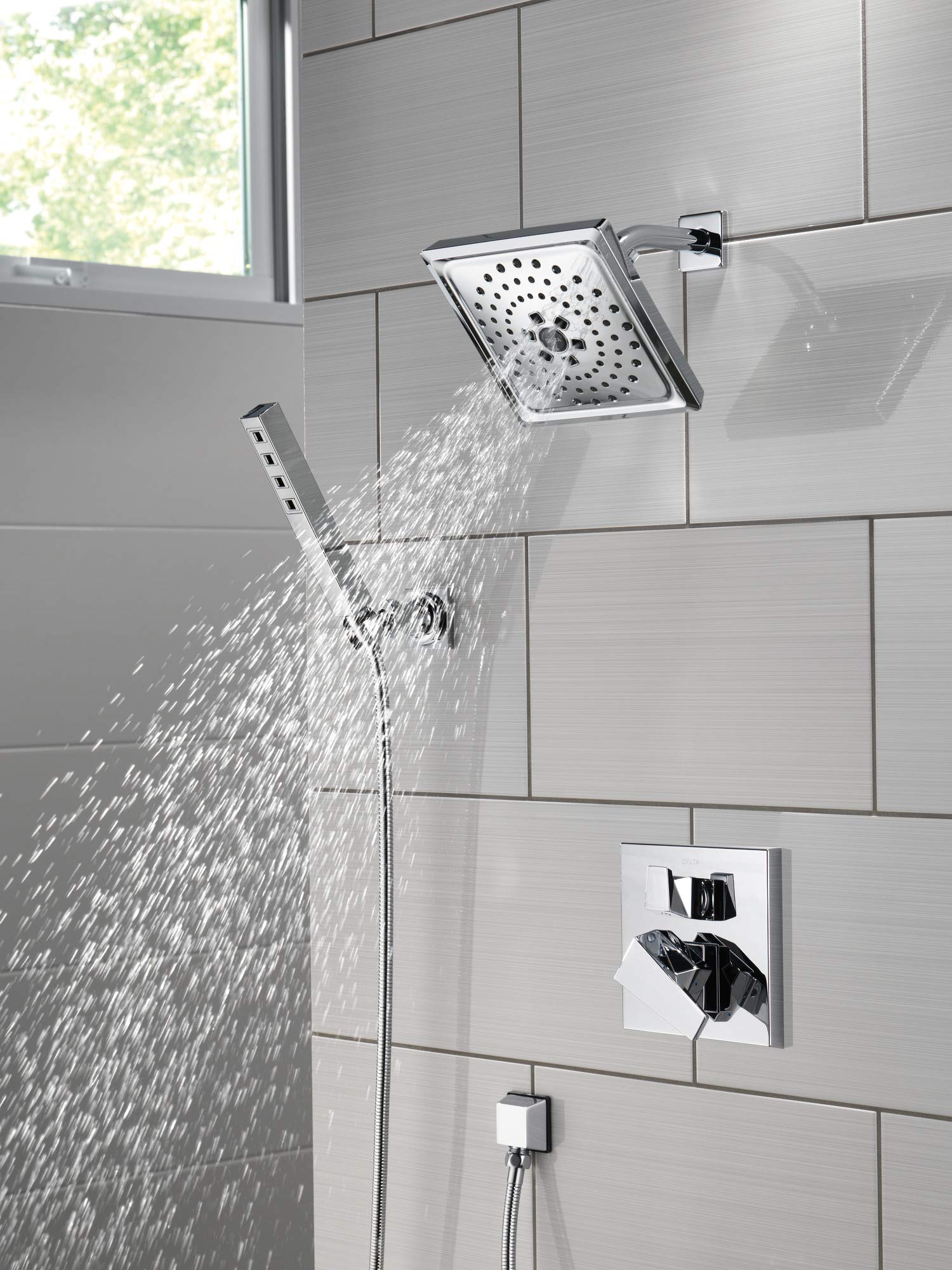 Black stainless steel shower arm with square rainfall head and handheld wand spraying water against tiled bathroom wall.