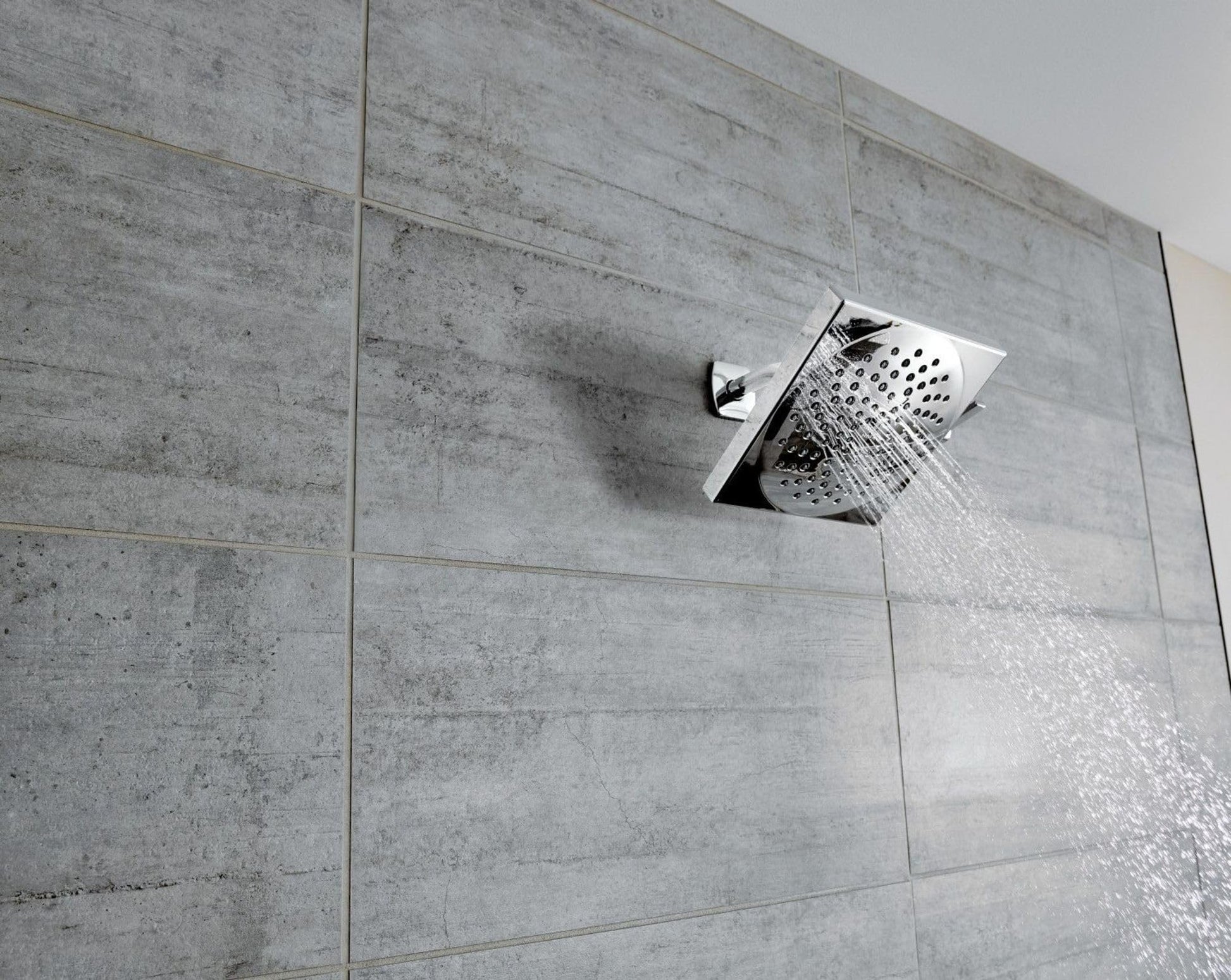 Chrome square rainshower showerhead mounted on gray tile wall with water spray visible.
