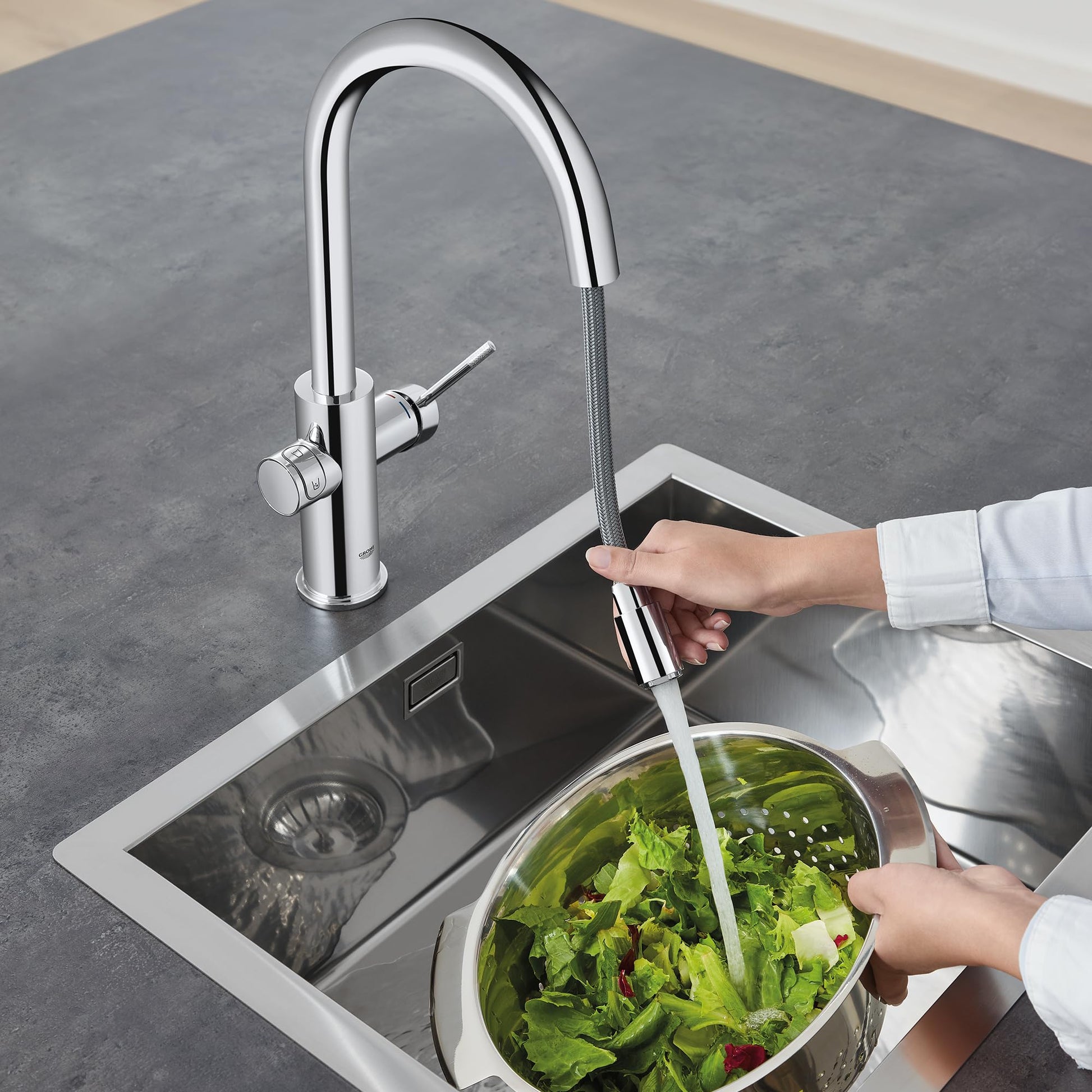 Chrome pull-down kitchen faucet with curved spout and single handle, shown dispensing water into a colander of lettuce above 