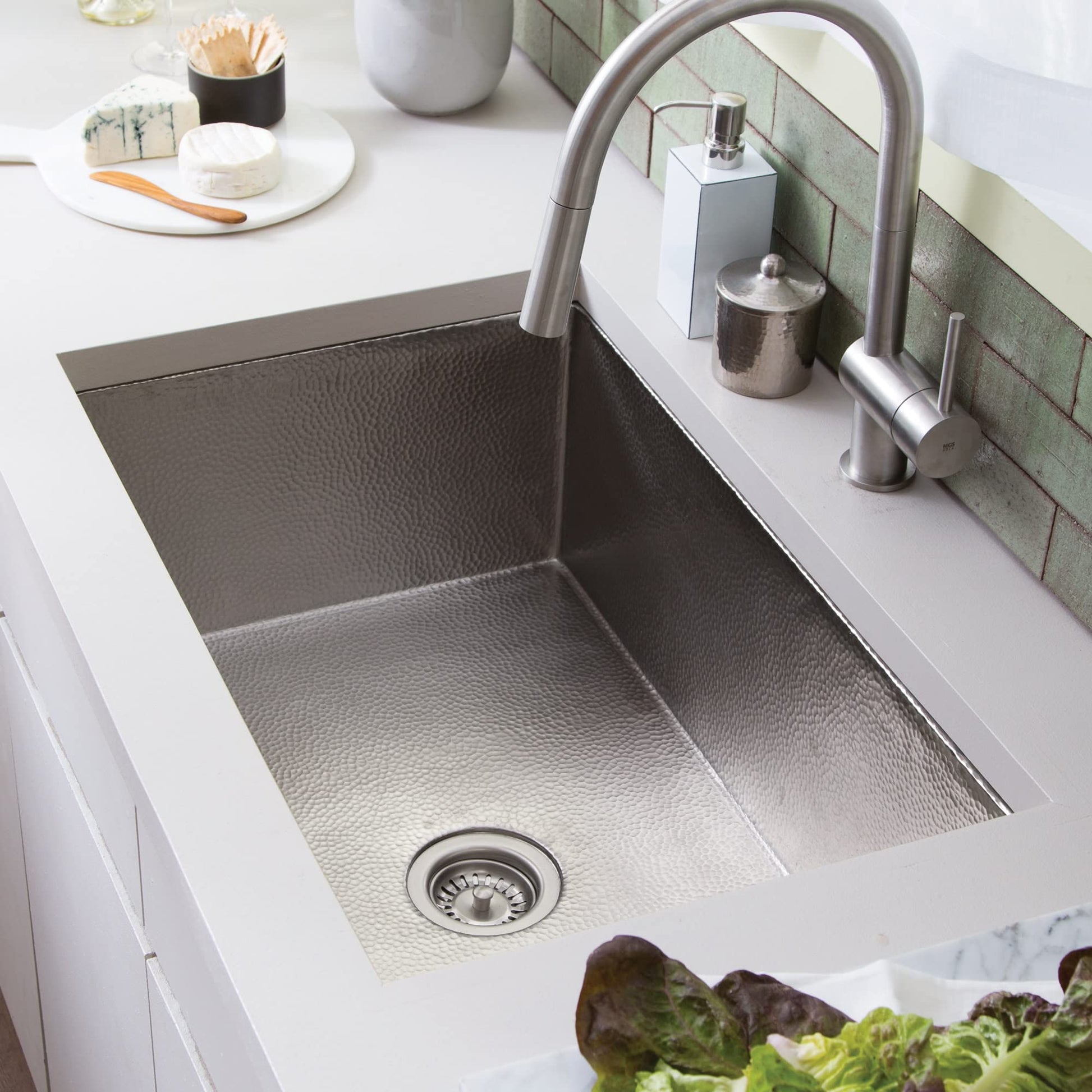 Brushed nickel undermount kitchen sink with hammered finish and arched faucet installed in white countertop.