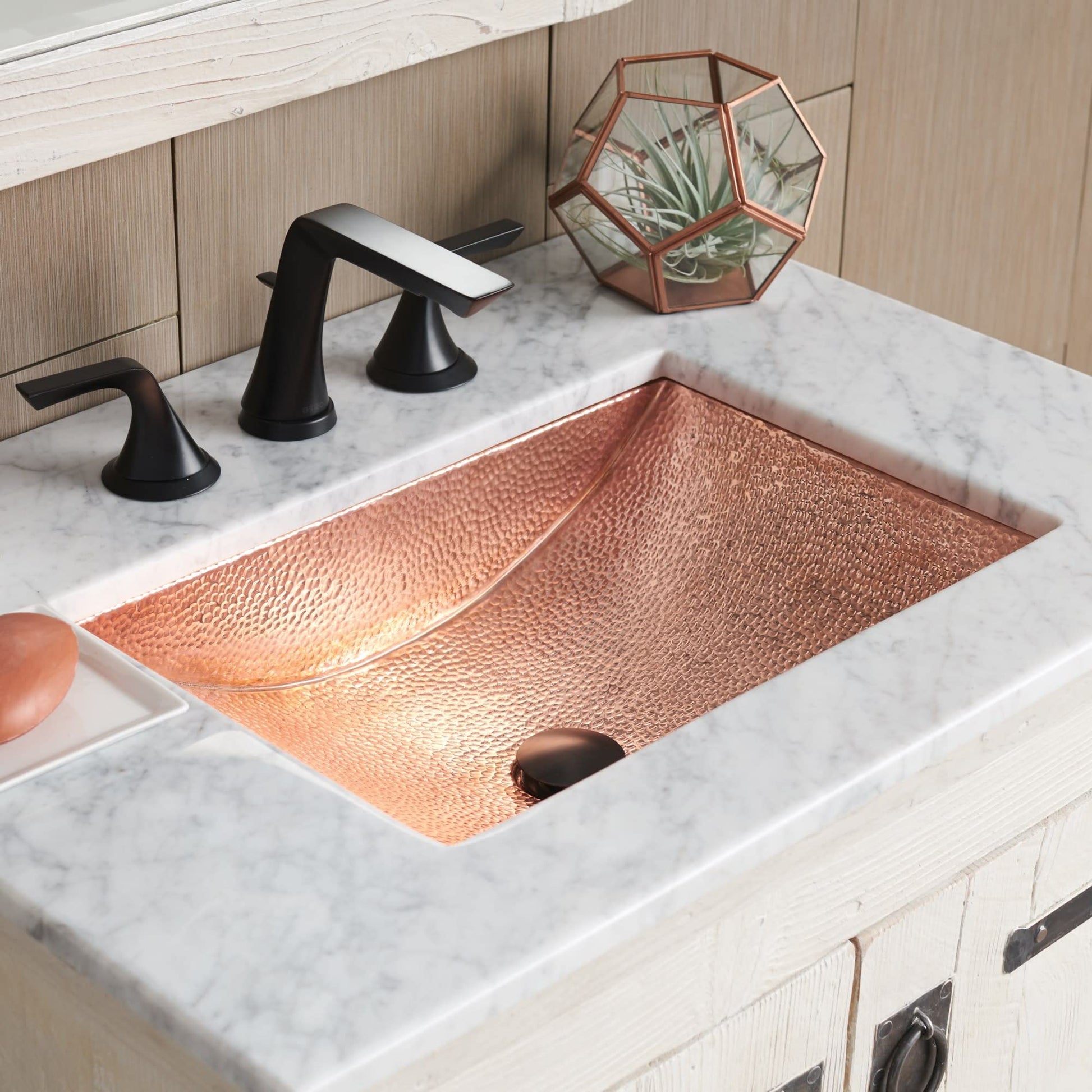 Polished copper rectangular sink with hammered texture surface mounted in white marble countertop with black faucet.