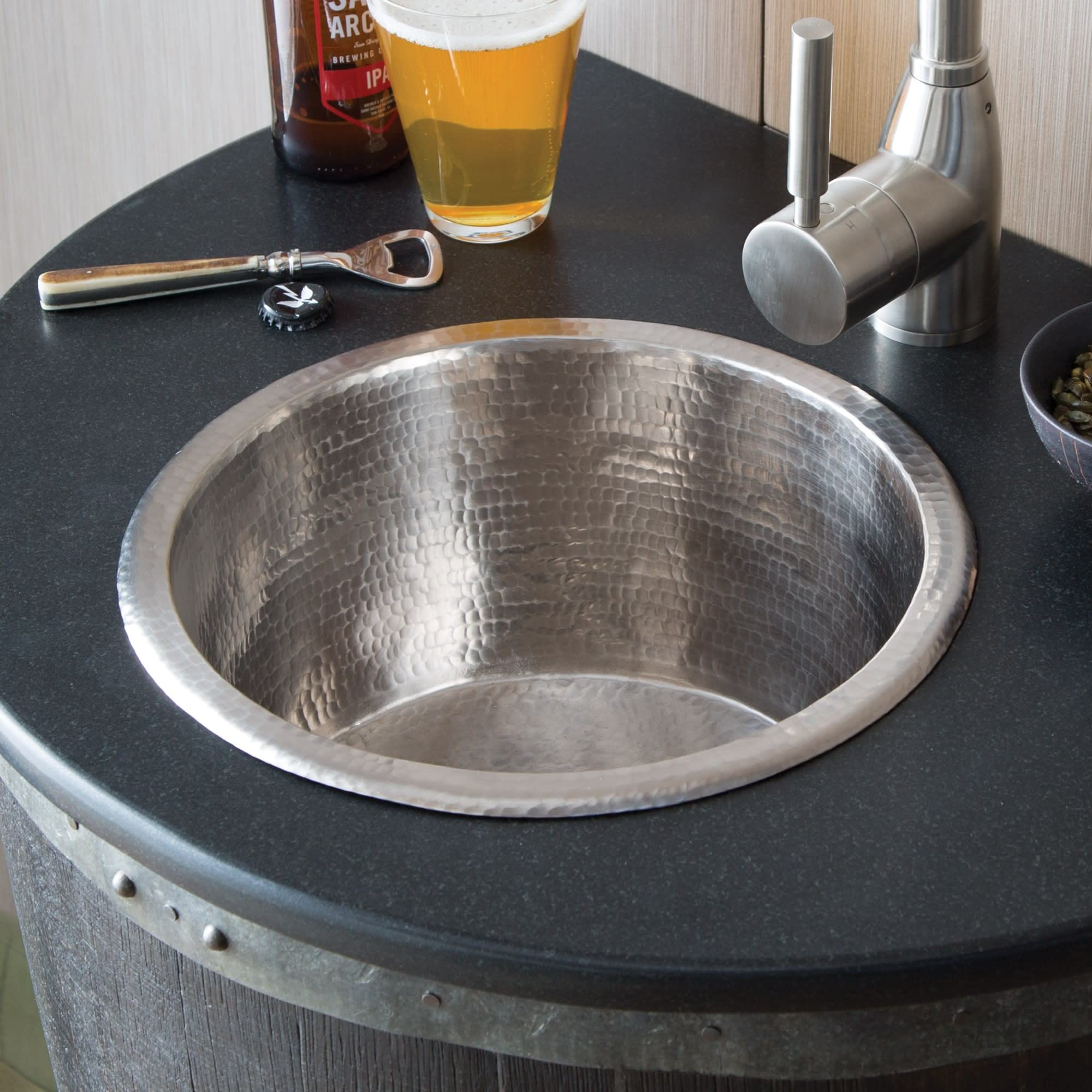 Hammered copper round bar sink with textured interior surface mounted in dark countertop, styled with faucet and bar accessor