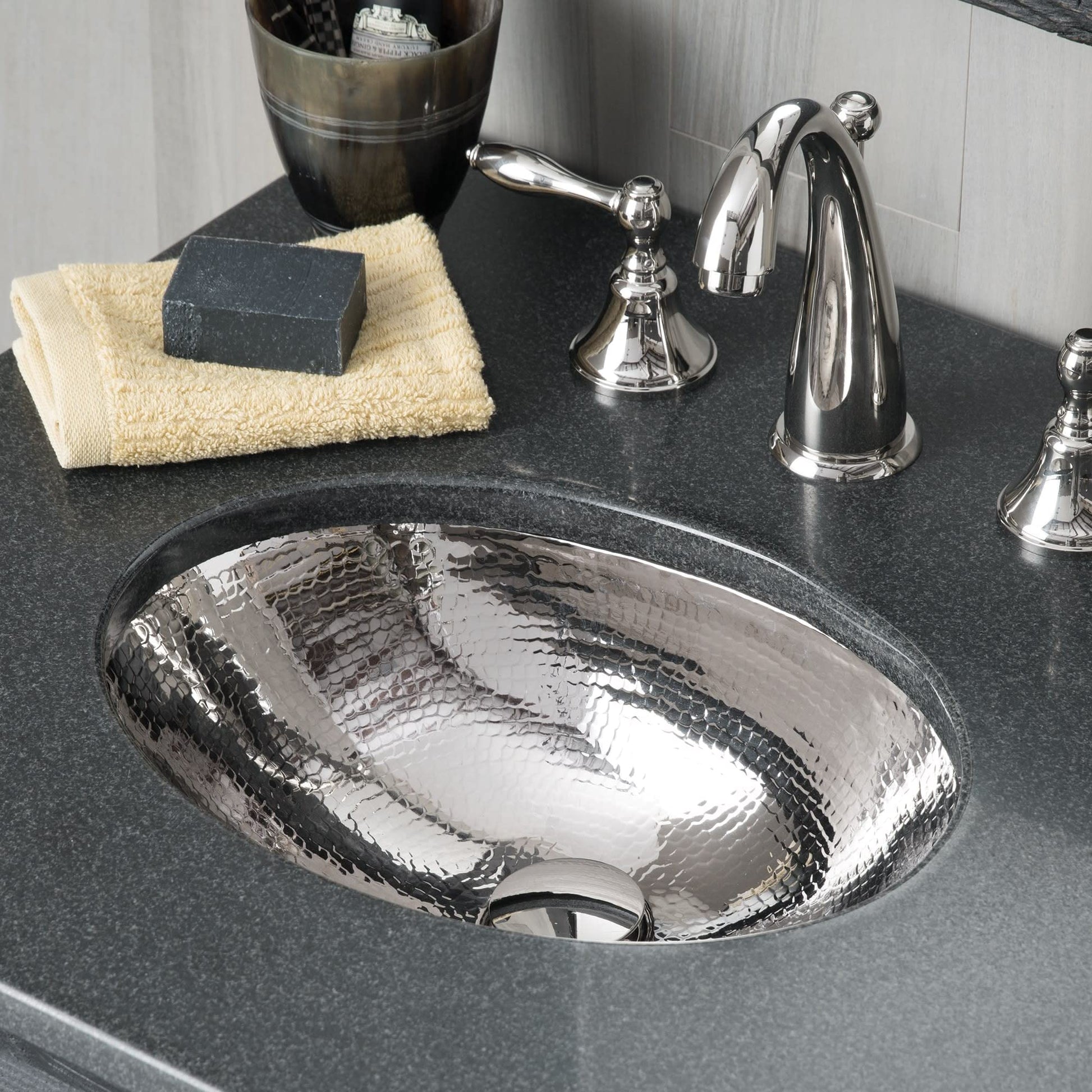 Hammered copper bathroom sink with organic oval shape mounted in dark countertop, chrome faucet and accessories nearby.