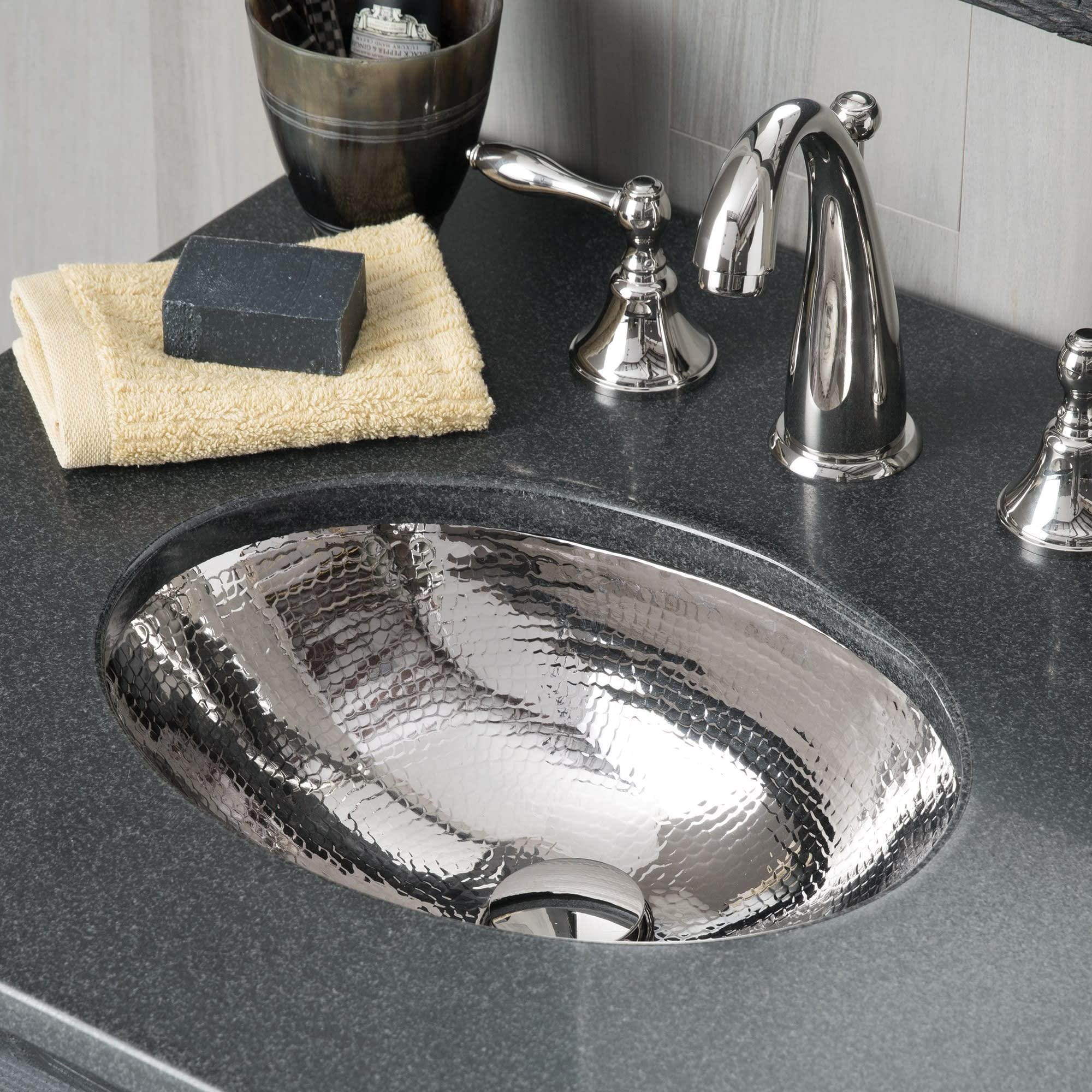 Polished nickel undermount sink with hammered texture displayed on dark countertop with faucet and bathroom accessories.