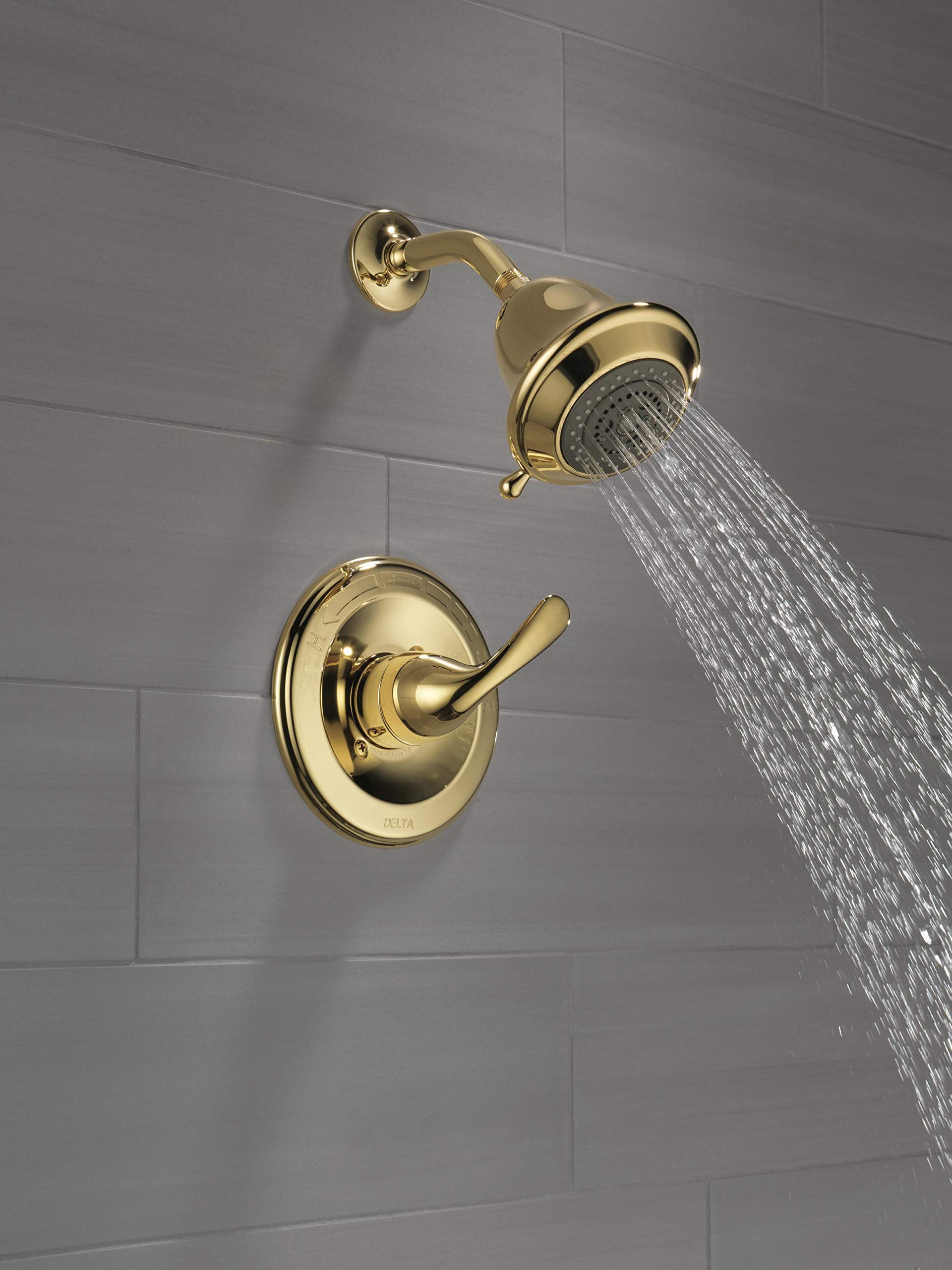 Polished brass shower trim with single lever handle and showerhead mounted on gray tile wall, water flowing from showerhead.