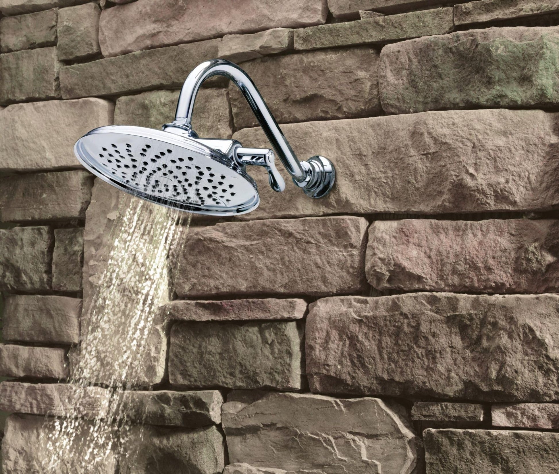 Chrome showerhead with oval faceplate and multiple spray holes mounted on curved arm against stone wall with water flowing.