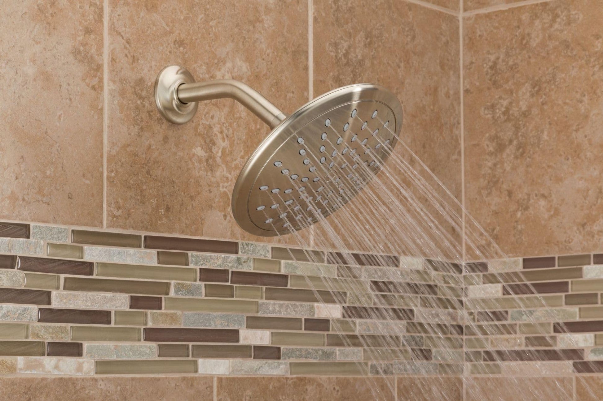 Stainless steel round shower head mounted on wall with curved arm, water spraying downward in bathroom with tan tile.