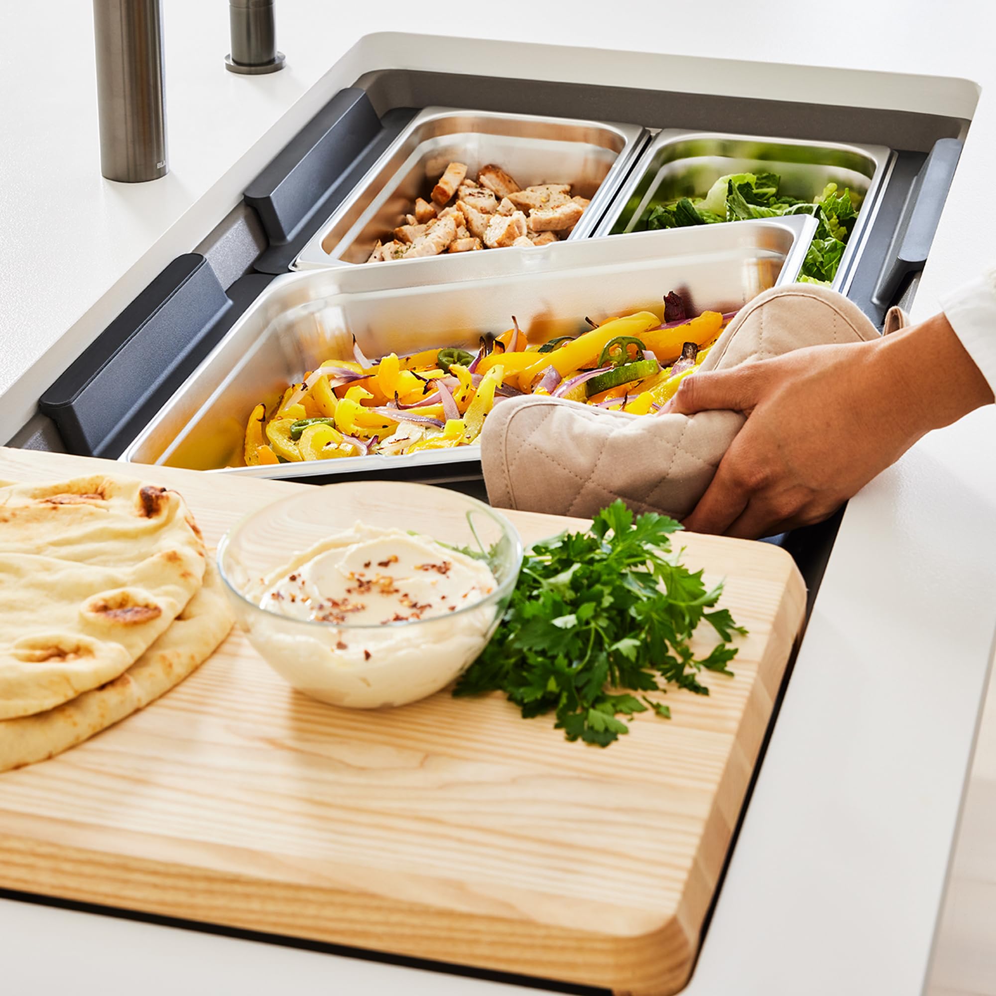 Stainless steel sink tray with multiple food containers holding cooked vegetables, grains, and greens, positioned next to a w
