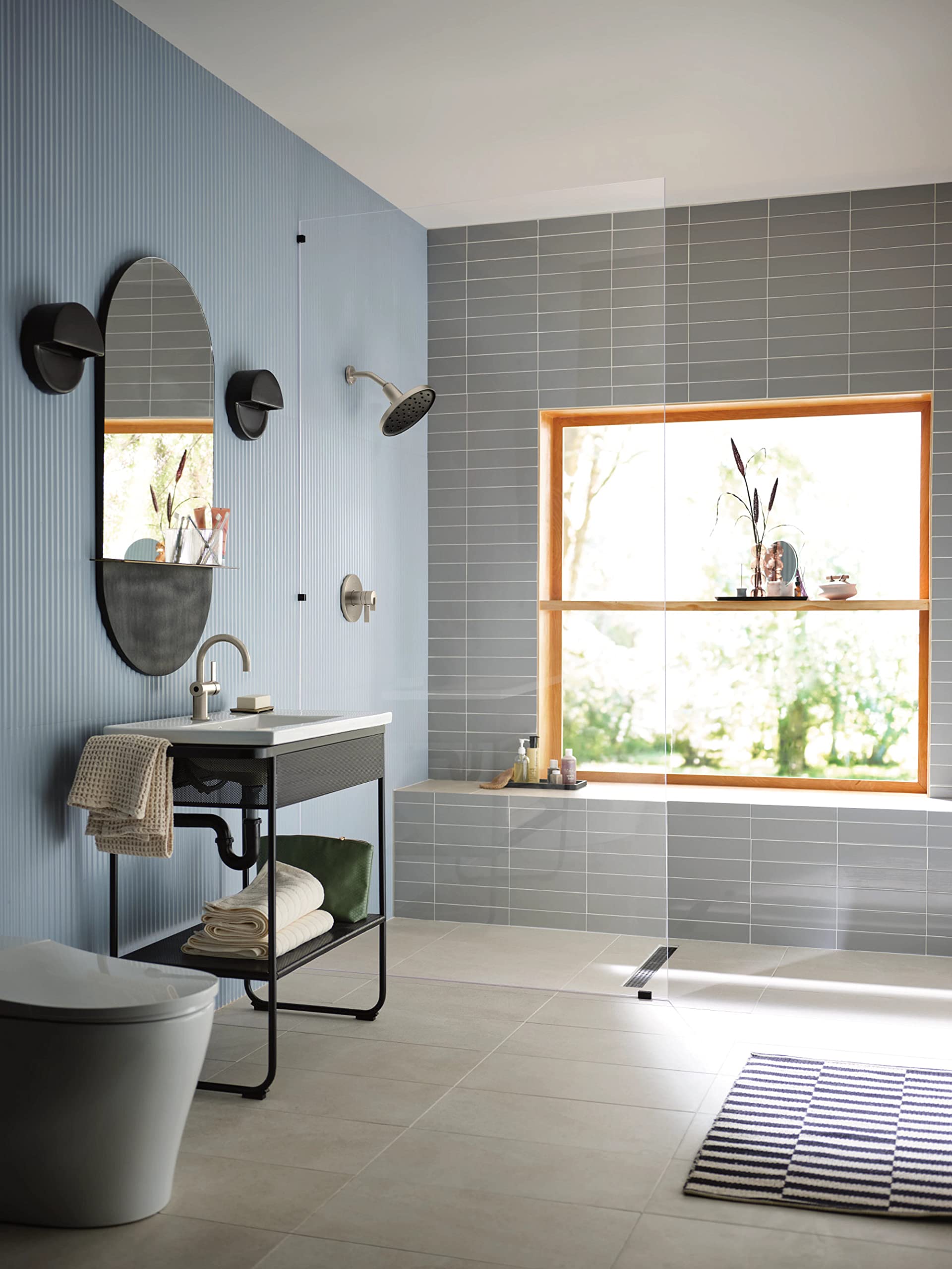 Round brushed nickel showerhead mounted on gray tile wall above bathtub in modern bathroom.