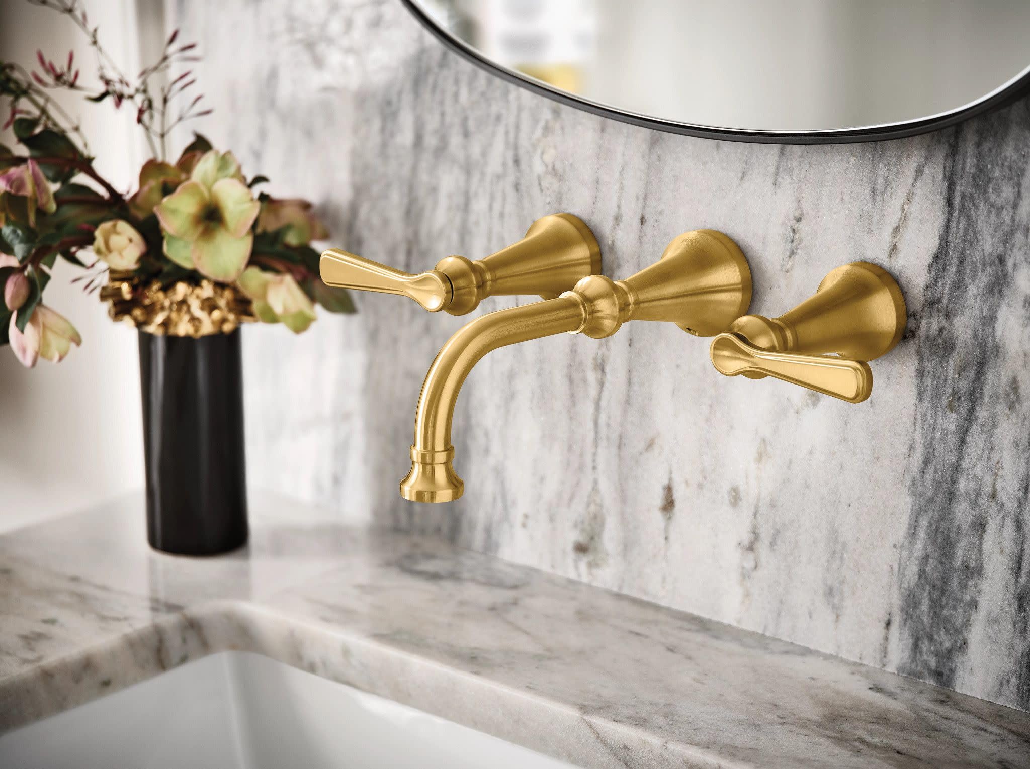 Brushed gold wall-mounted bathroom faucet with two lever handles and curved spout installed above marble sink.