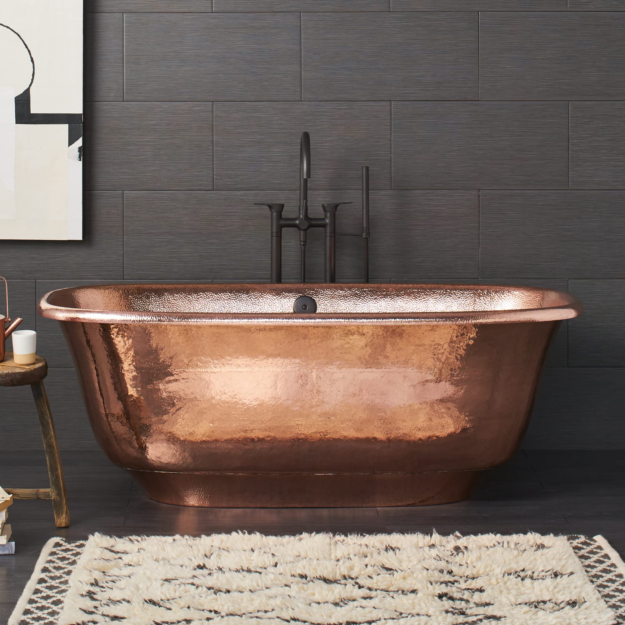 Antique copper bathtub with hammered finish and sloped sides against dark tile wall with black faucet.