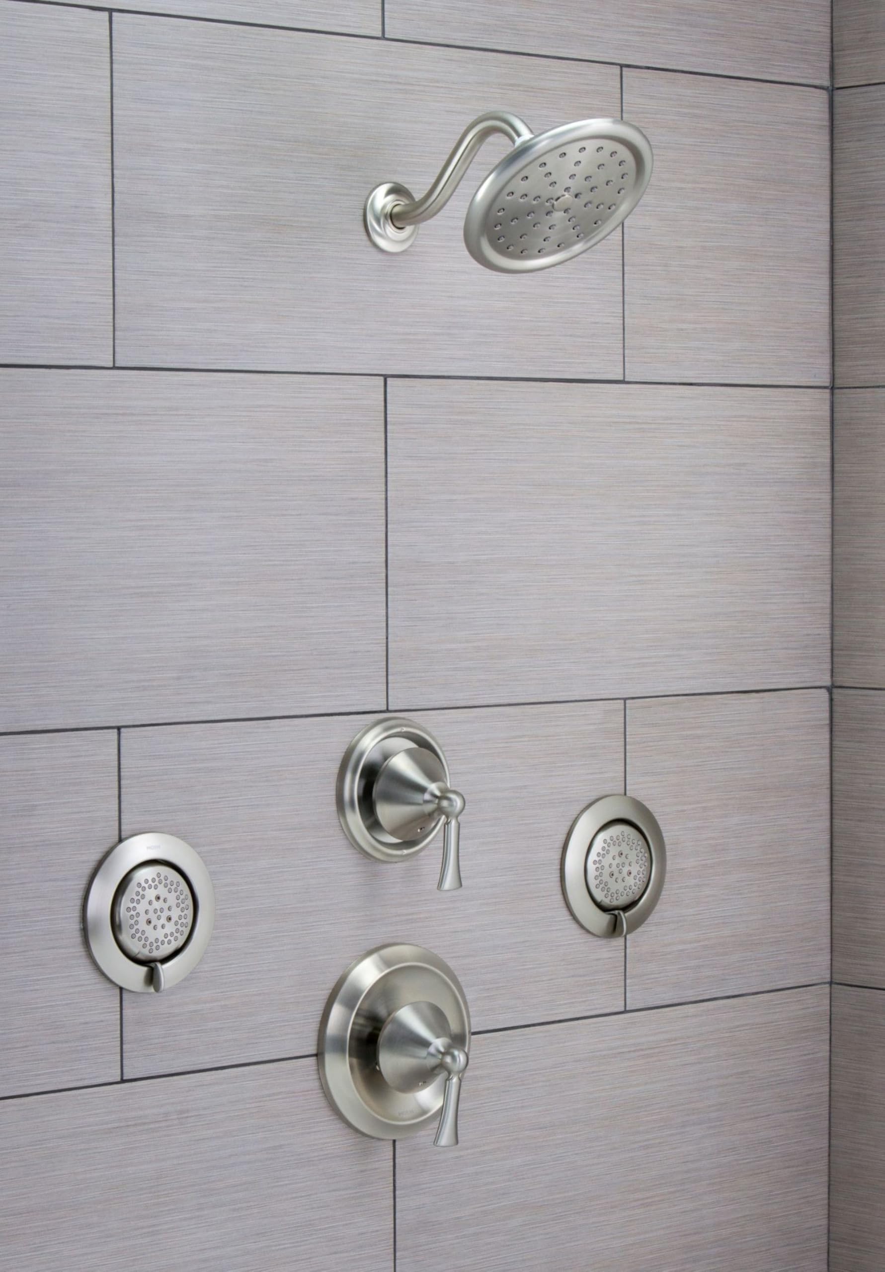 Brushed nickel shower head spray with perforated oval face mounted on curved arm against tiled shower wall.