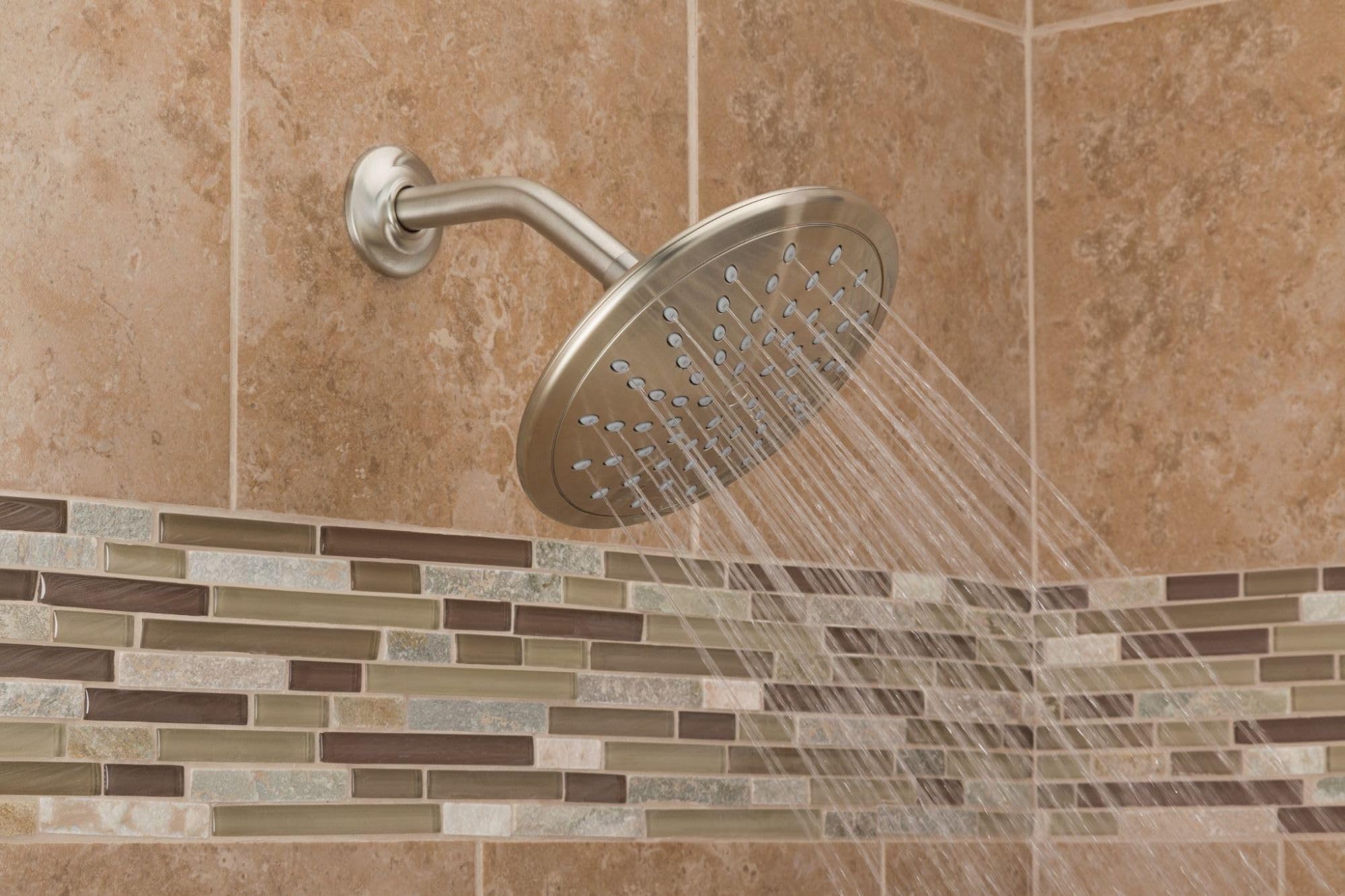 Brushed nickel circular rain shower head mounted on wall with curved arm, water spray visible, installed above tile backsplas