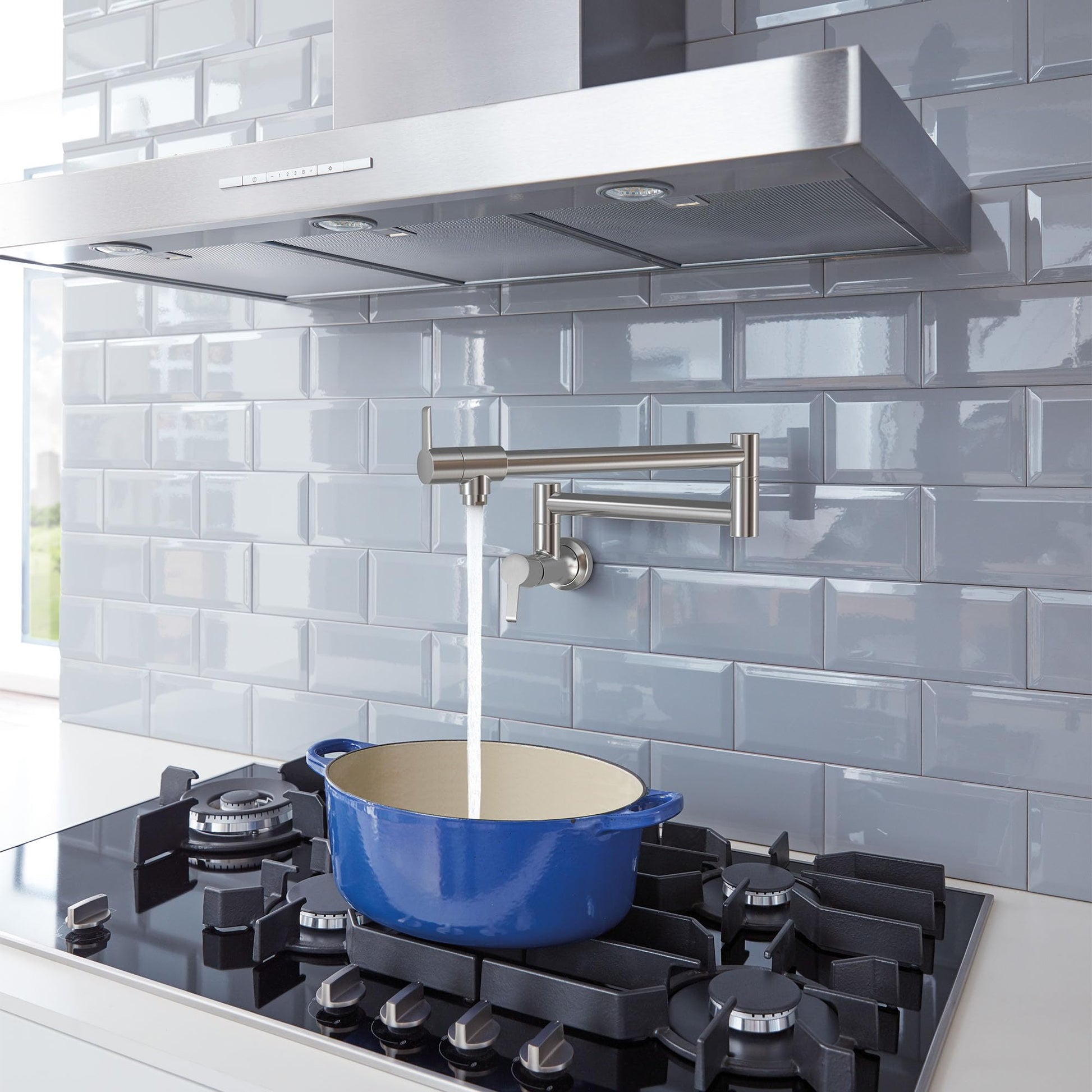 Stainless steel wall-mounted pot filler faucet with two handles positioned above a stovetop with a blue pot.