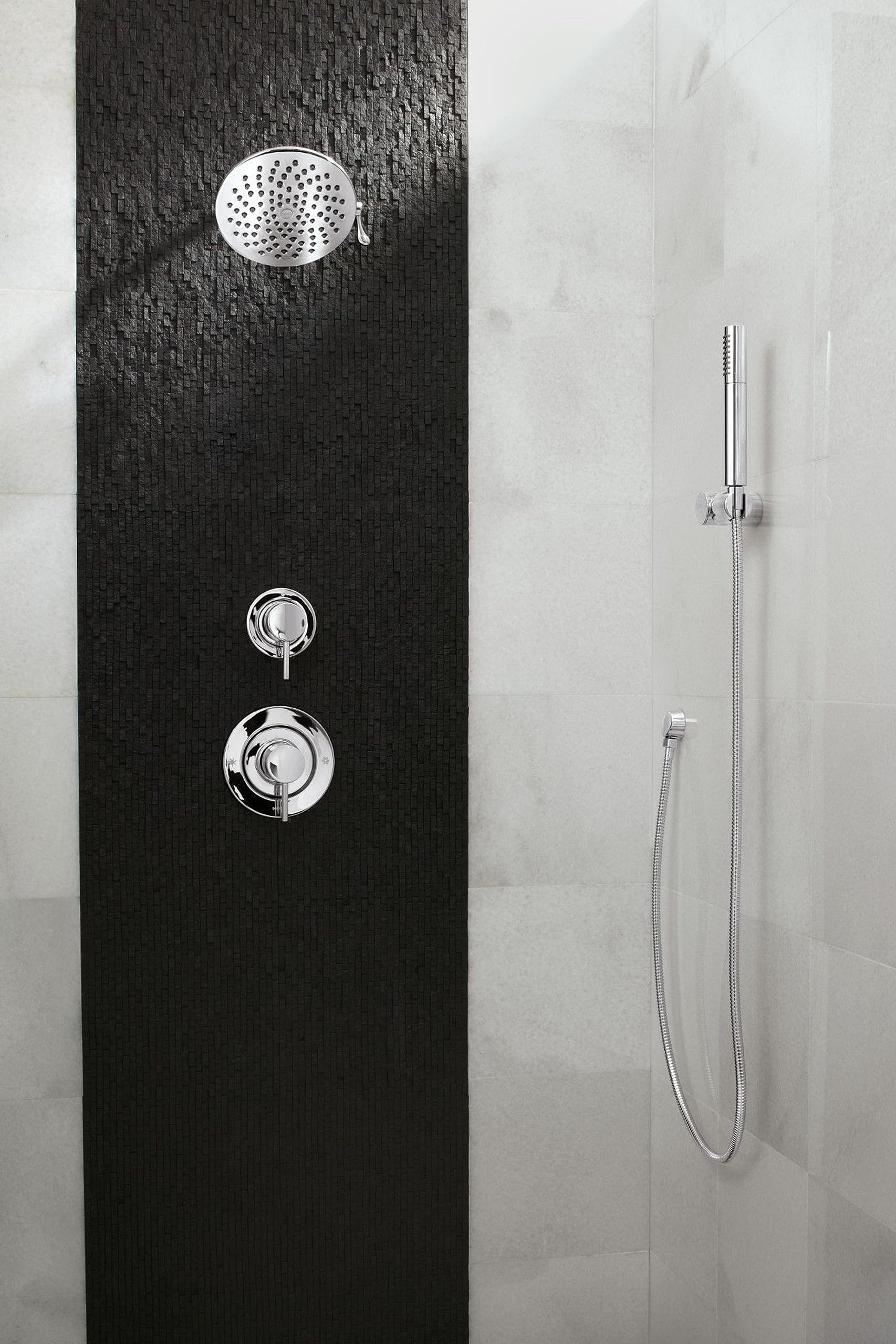Chrome shower valve trim with round showerhead, control handles, and handheld shower wand mounted on black and white tile wal