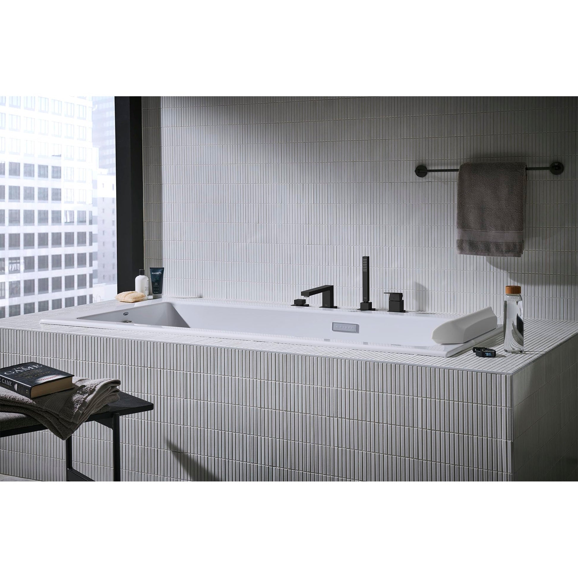 Matte black four-hole roman tub faucet with single handle and hand shower mounted on white tile deck surrounding a bathtub.