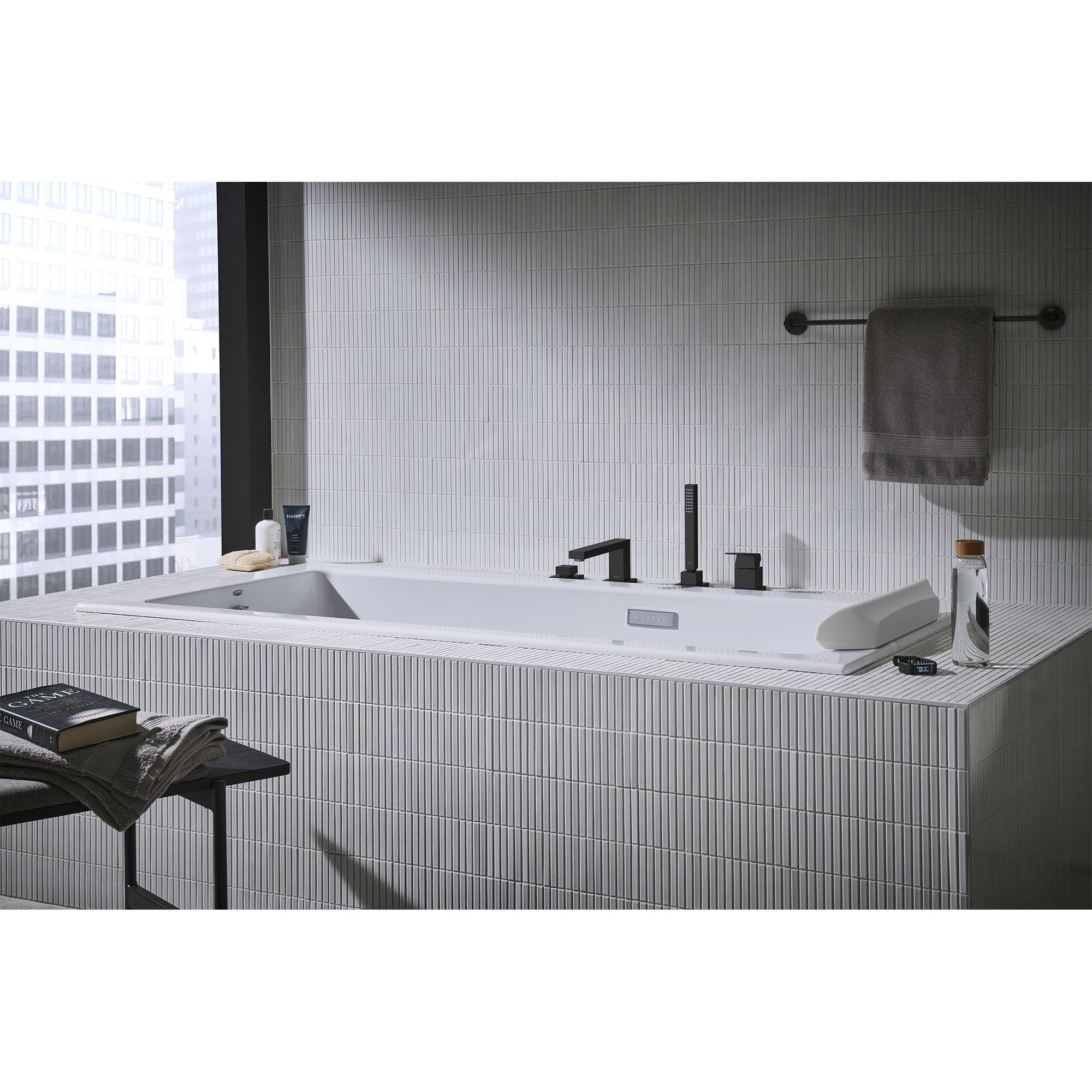 Matte black four-hole roman tub faucet with single handle and hand shower mounted on white tile deck surrounding a bathtub.