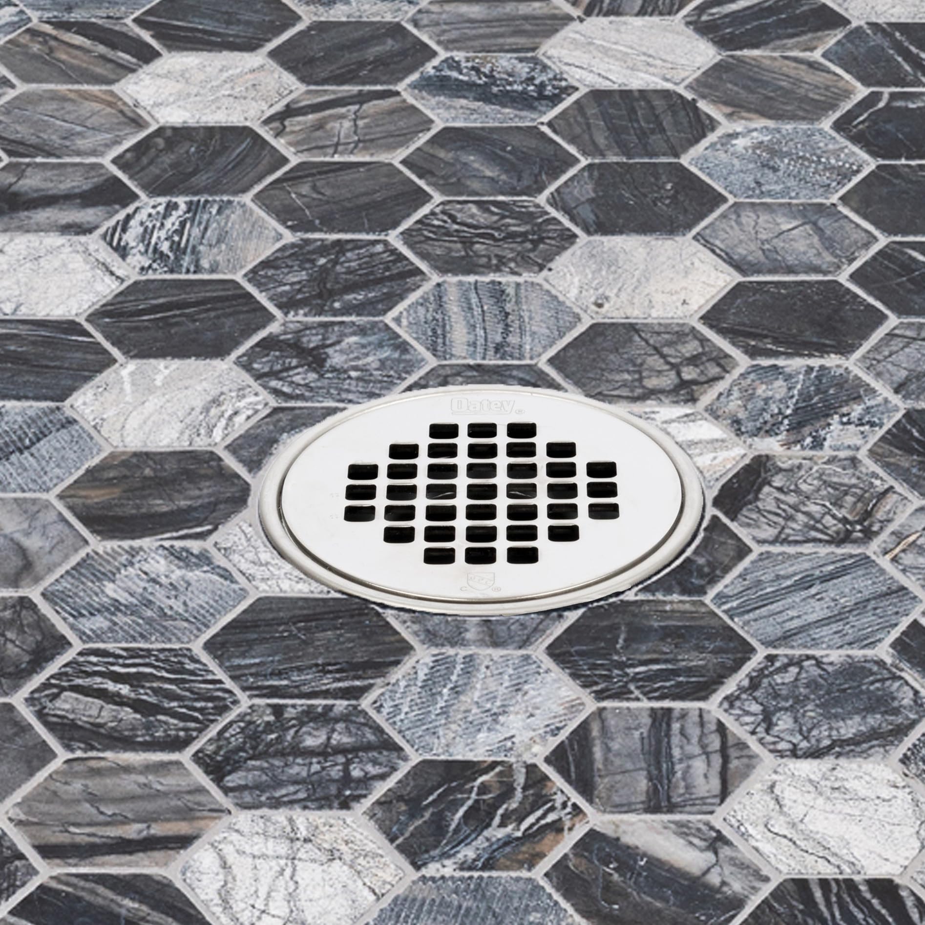 Satin nickel shower strainer with oval shape and rectangular drain holes installed on hexagonal marble tile floor.