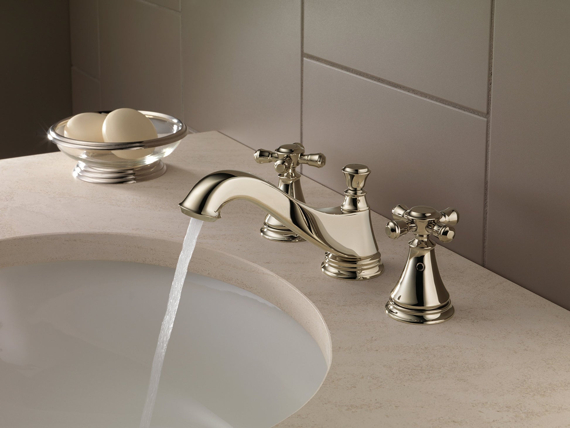Polished nickel three-piece bathroom faucet with curved spout and cross handles mounted on beige countertop above sink.