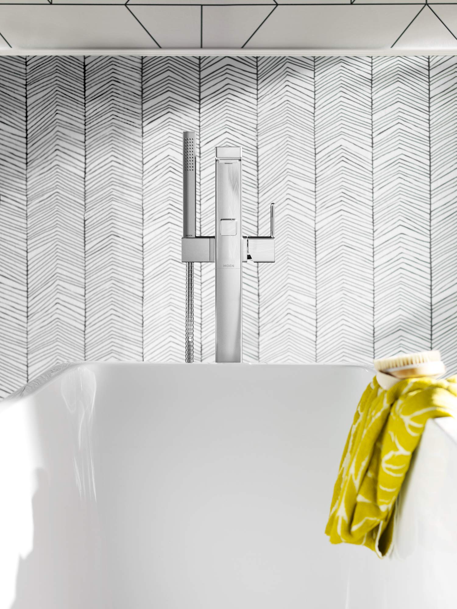 Chrome floor-mount tub filler with single handle, spout, and handheld showerhead mounted on herringbone tile wall above white
