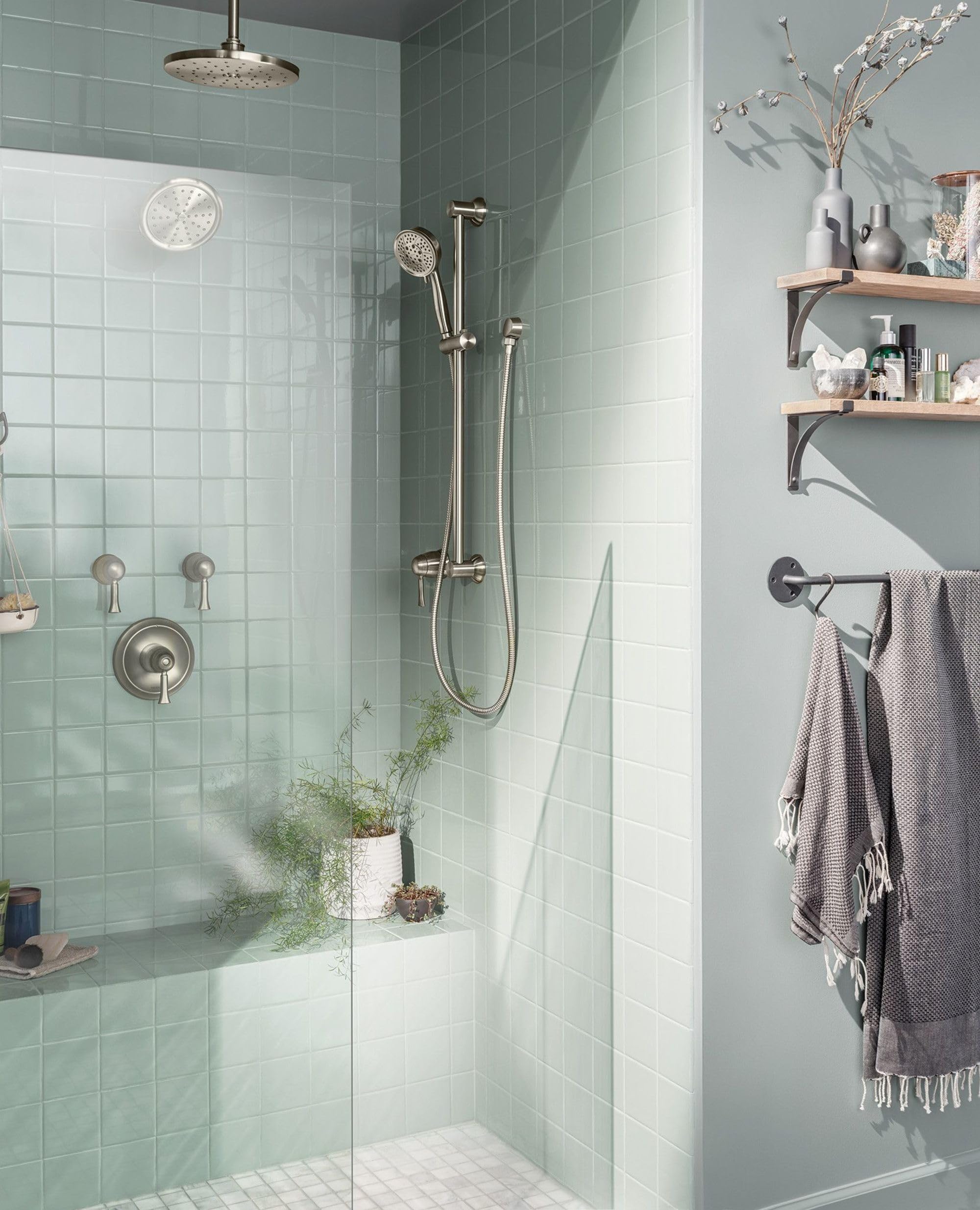 Brushed nickel shower arm flange mounted on tiled bathroom wall, connecting vertical pipe to overhead showerhead.