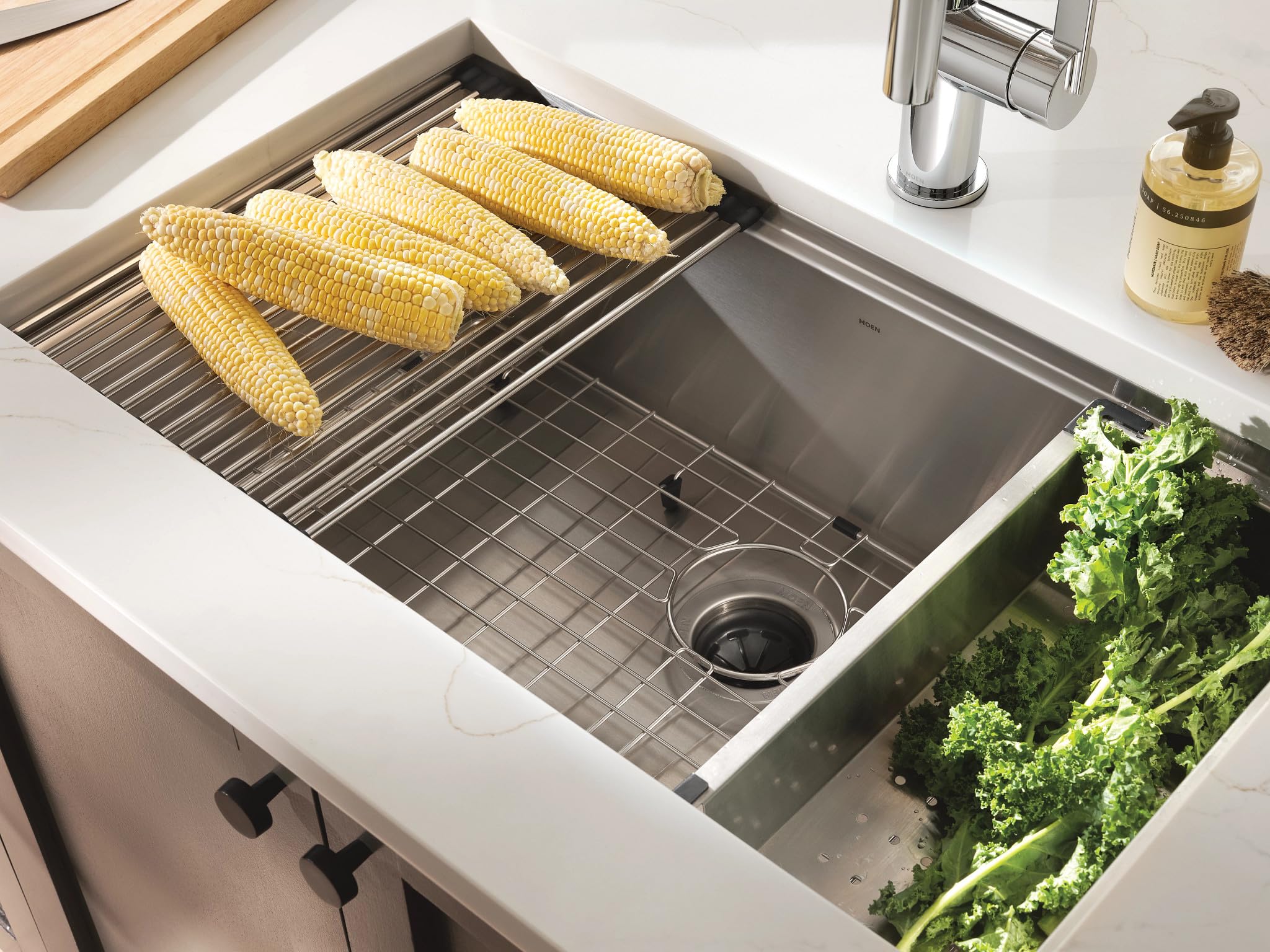 Stainless steel undermount sink with drying rack holding corn, grid insert, colander compartment, and faucet visible.