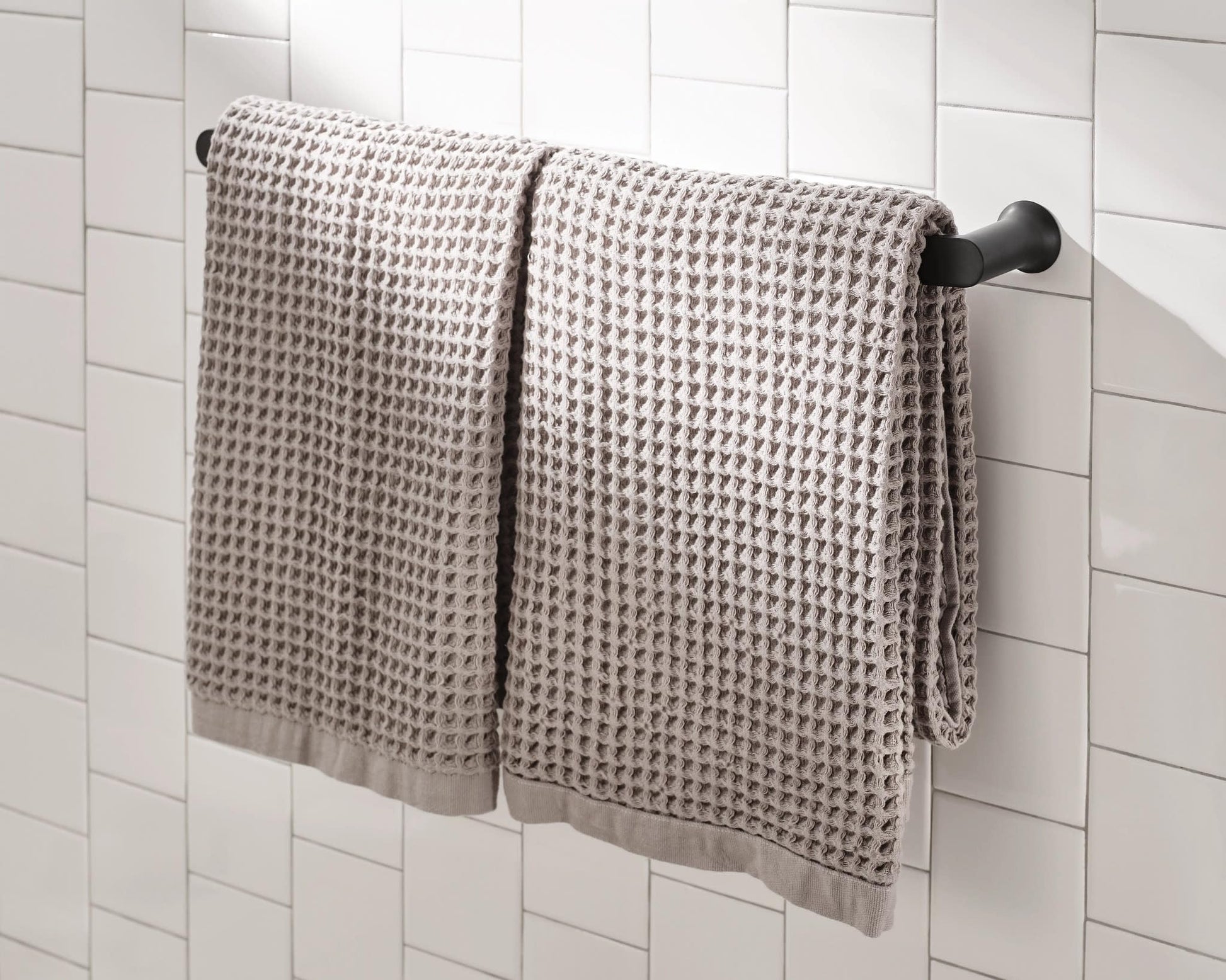 Black wall-mounted towel bar holding two gray textured towels against white subway tile.
