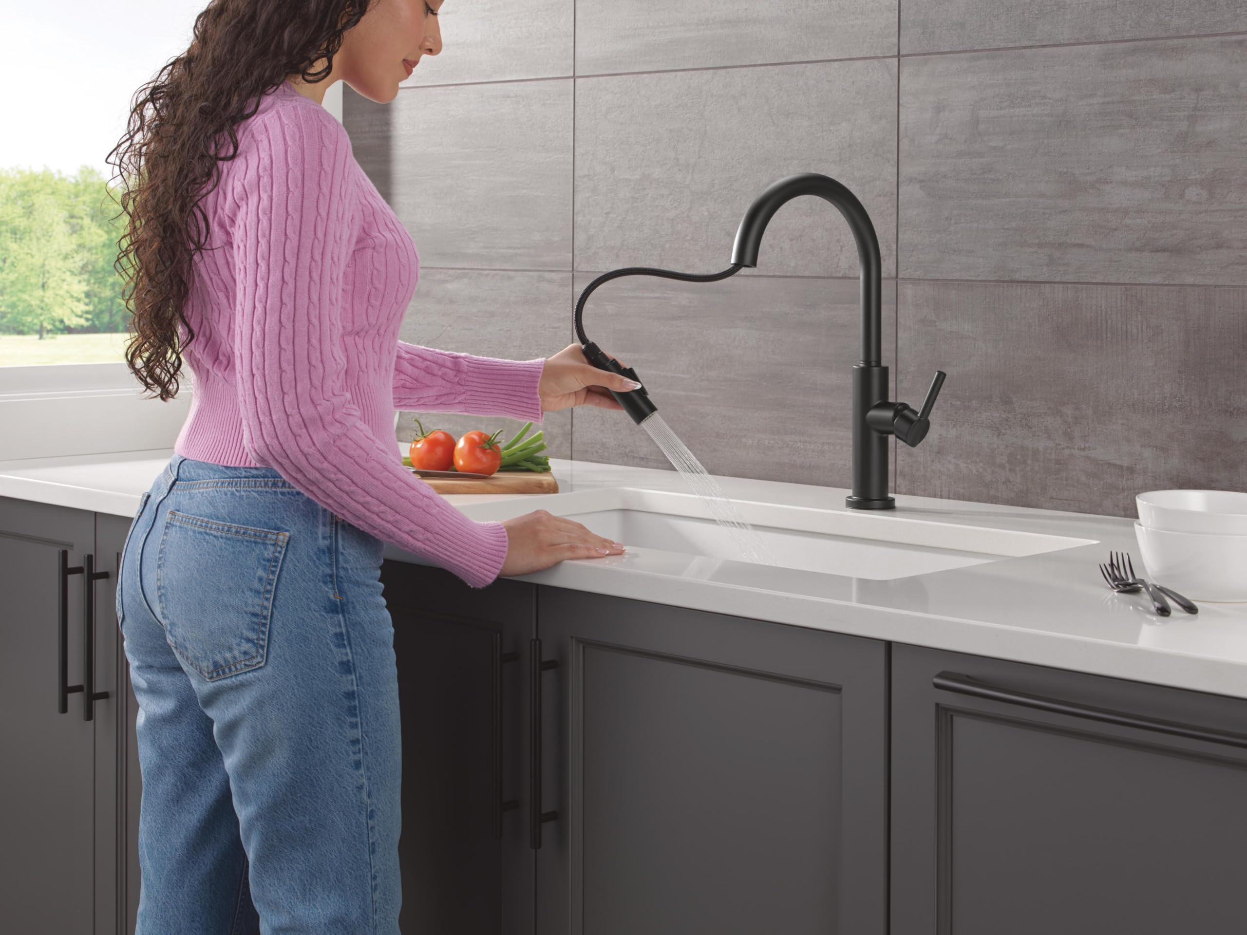 Matte black single-handle pull-down kitchen faucet with curved spout installed above white sink in gray tile kitchen.