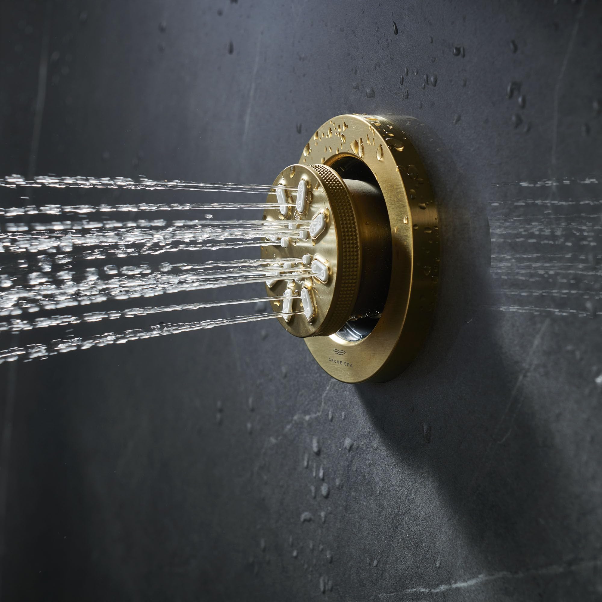 Round brushed gold body spray mounted on gray tile wall with two water spray streams visible.