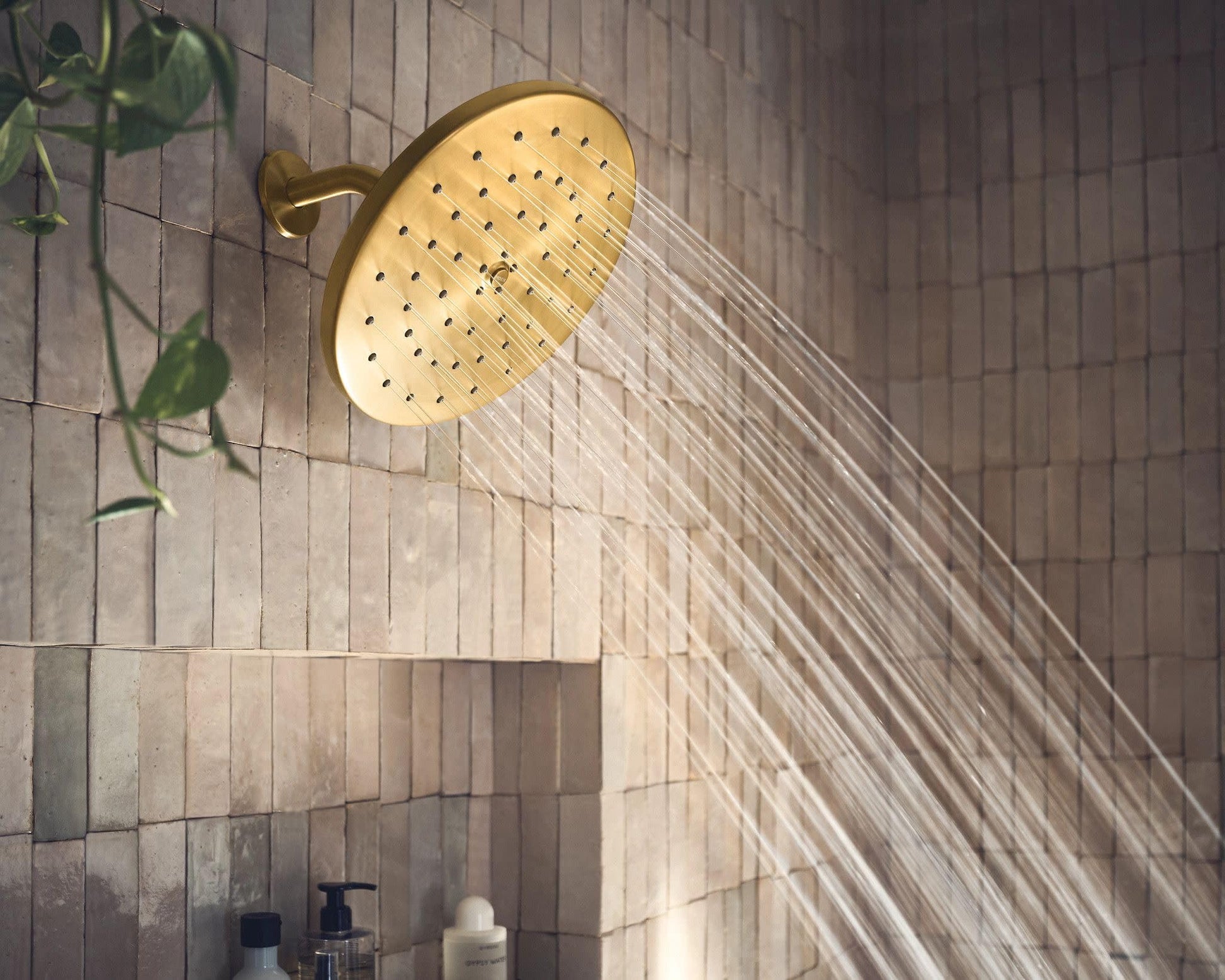 Brushed gold circular showerhead with multiple spray holes mounted on tiled bathroom wall with water stream visible.