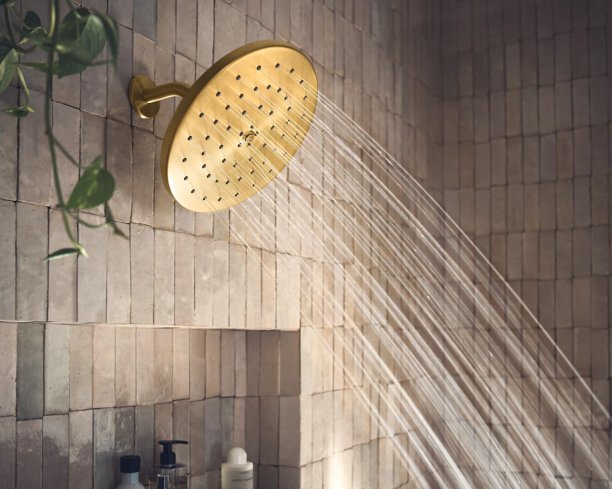 Brushed gold circular showerhead with multiple spray holes mounted on tiled bathroom wall with water stream visible.
