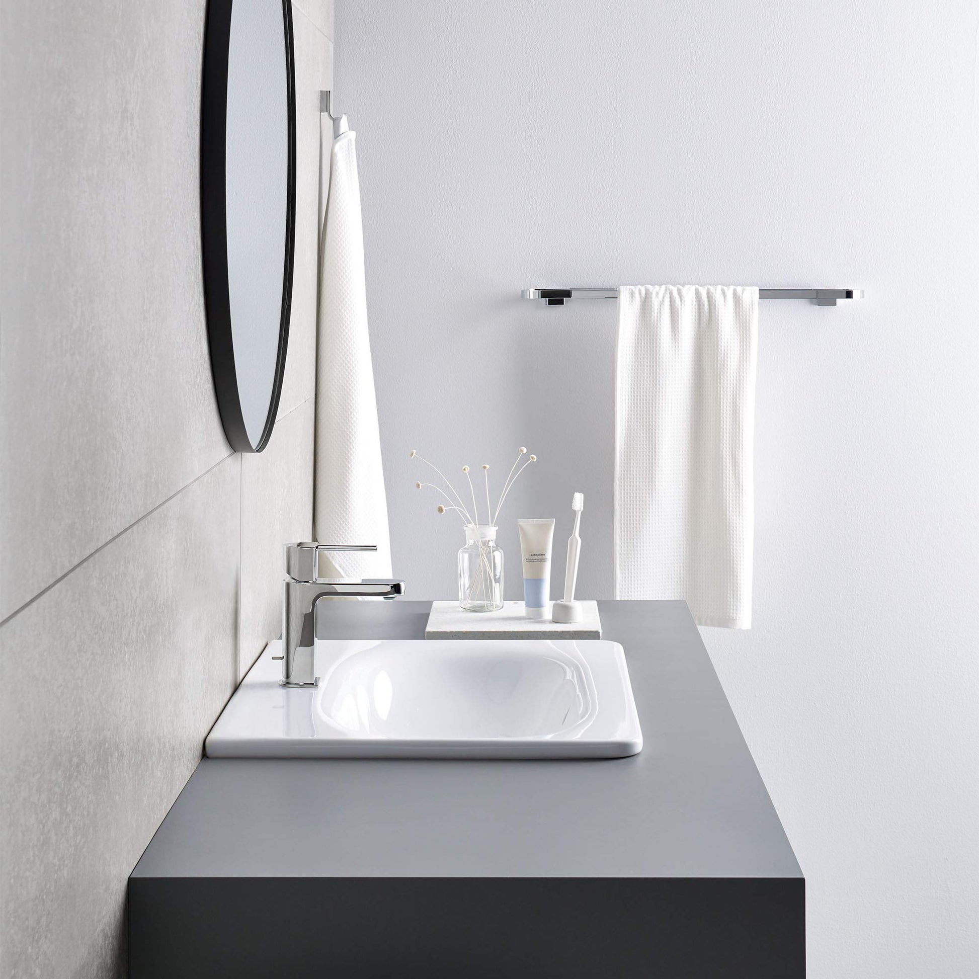 Chrome towel bar mounted on white bathroom wall above sink with white towel draped over it.