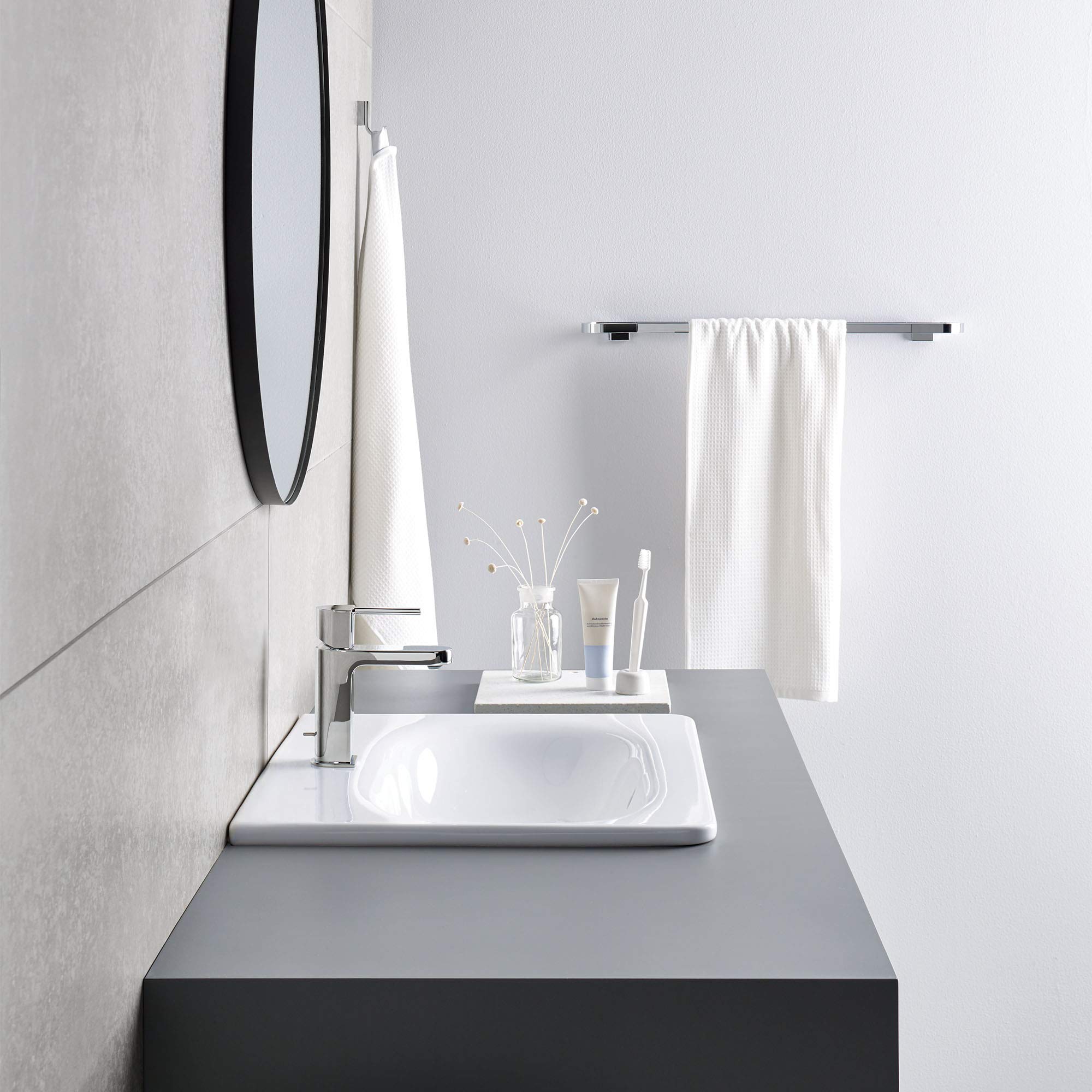 Chrome towel bar mounted on white bathroom wall above sink with white towel draped over it.