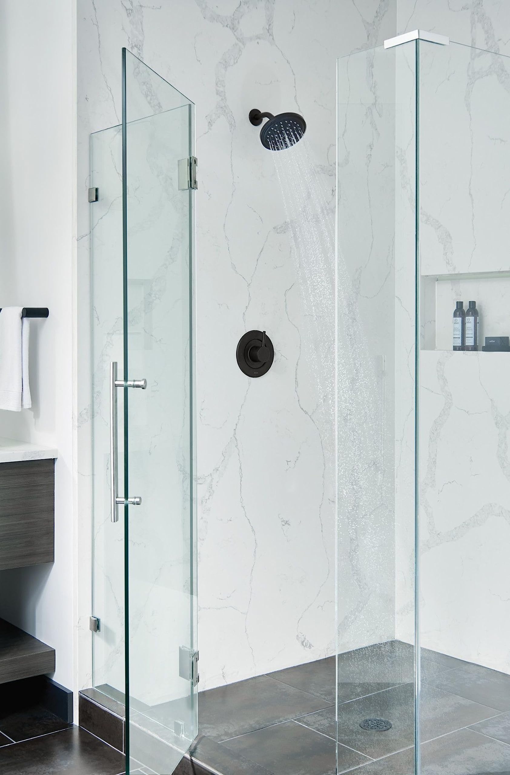 Matte black circular shower head mounted on marble tile wall with water streaming downward in a shower enclosure.
