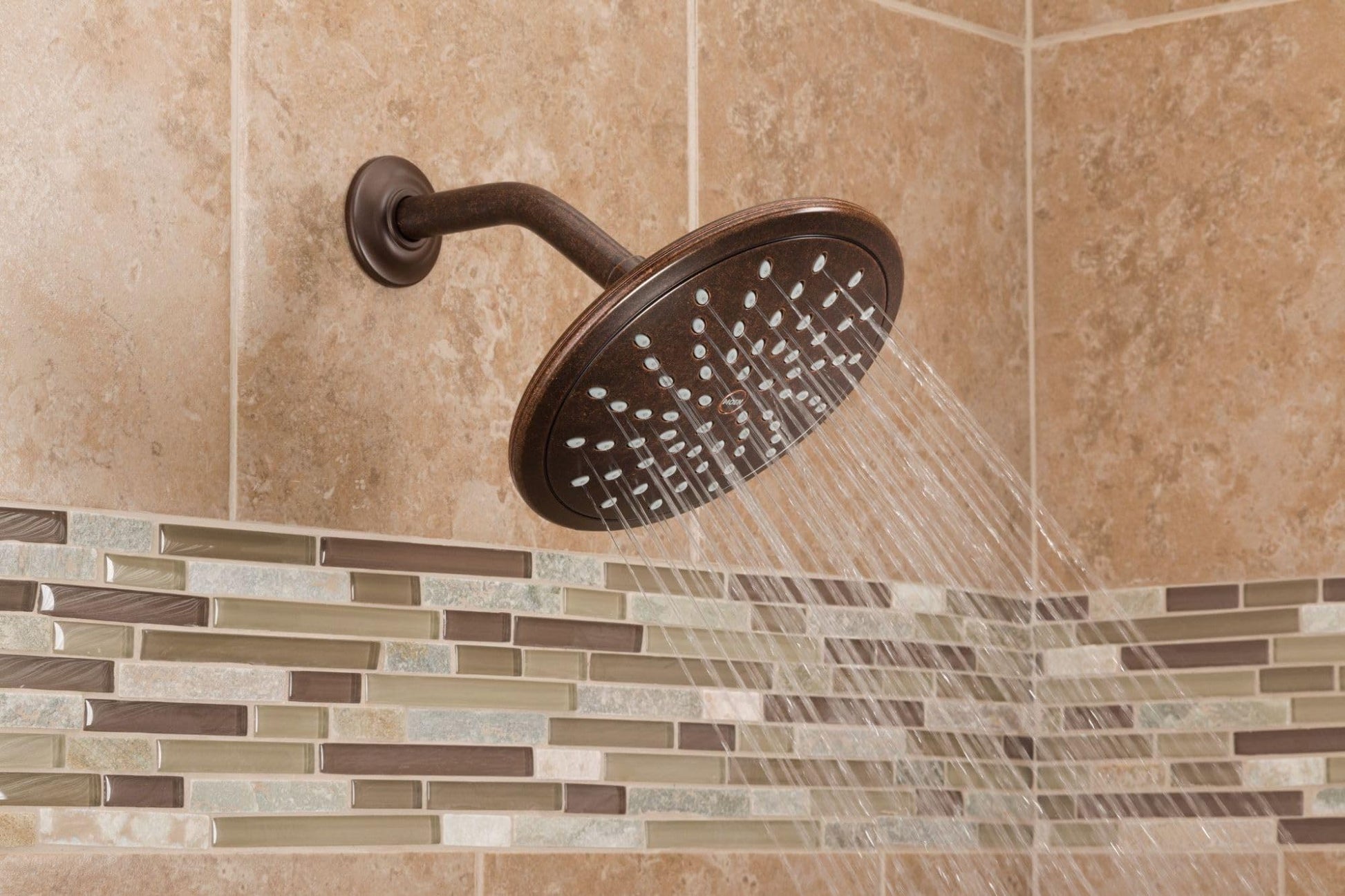 Brushed nickel round rain shower head mounted on wall with curved arm, water streaming downward over tiled bathroom wall.