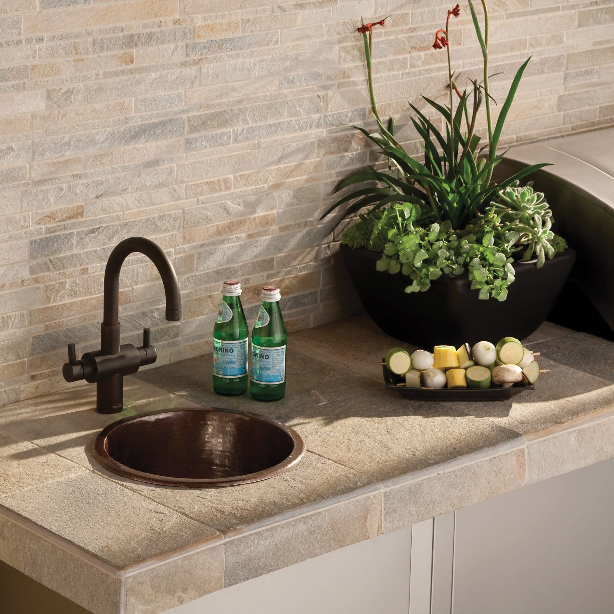 Hammered copper bar sink with dark bronze faucet installed in beige stone countertop.