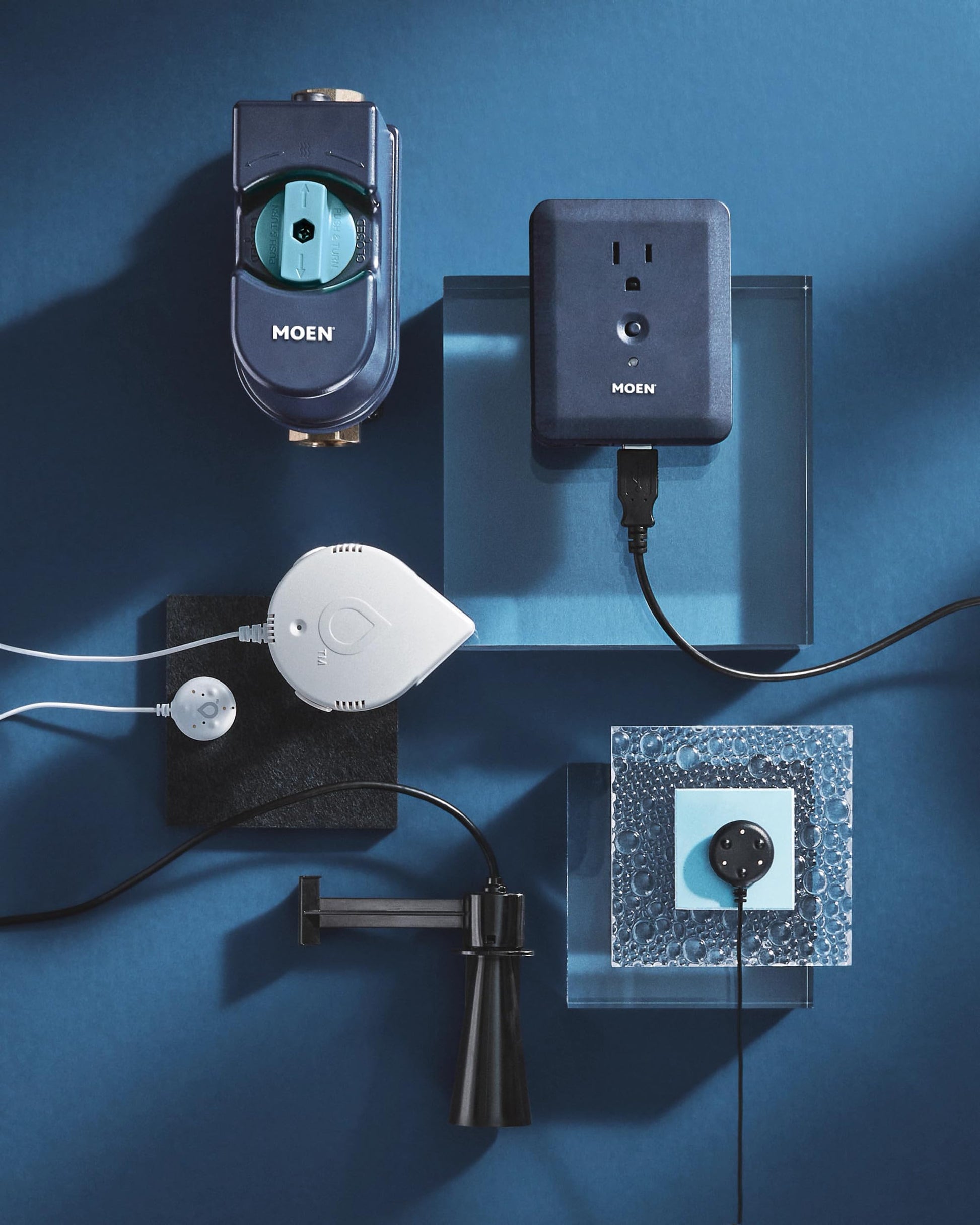 Moen Flo smart water monitor system components including black shutoff valve, power adapter, white sensor hub, and extension 