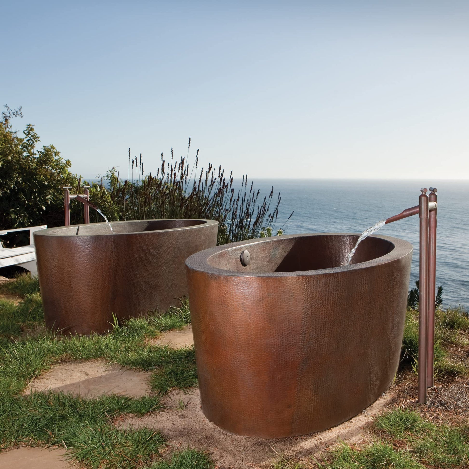 Hammered copper freestanding bathtub with textured surface and sloped interior, displayed outdoors near ocean.