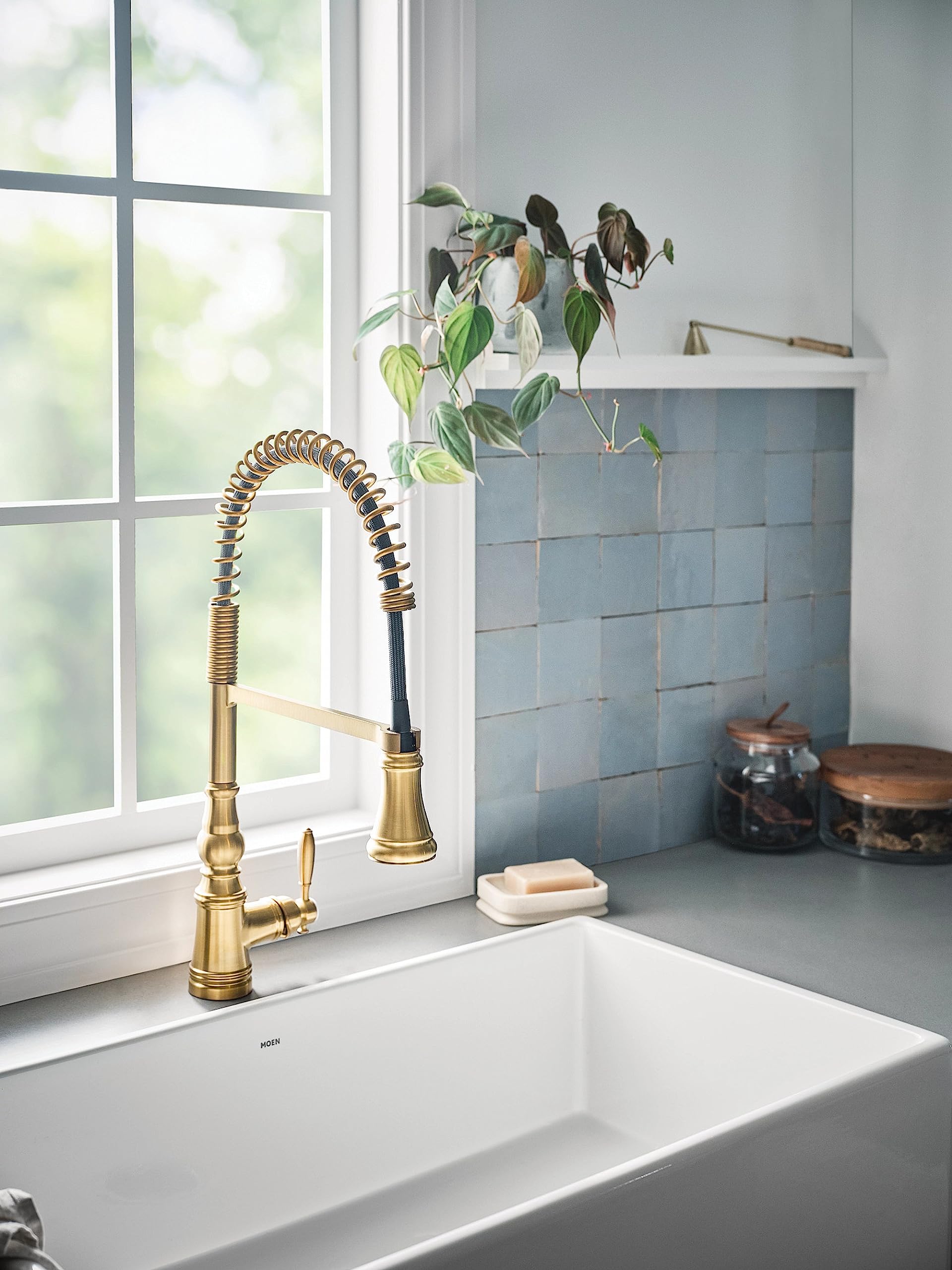 Brushed gold pulldown kitchen faucet with coiled spring hose and single handle mounted above white farmhouse sink.