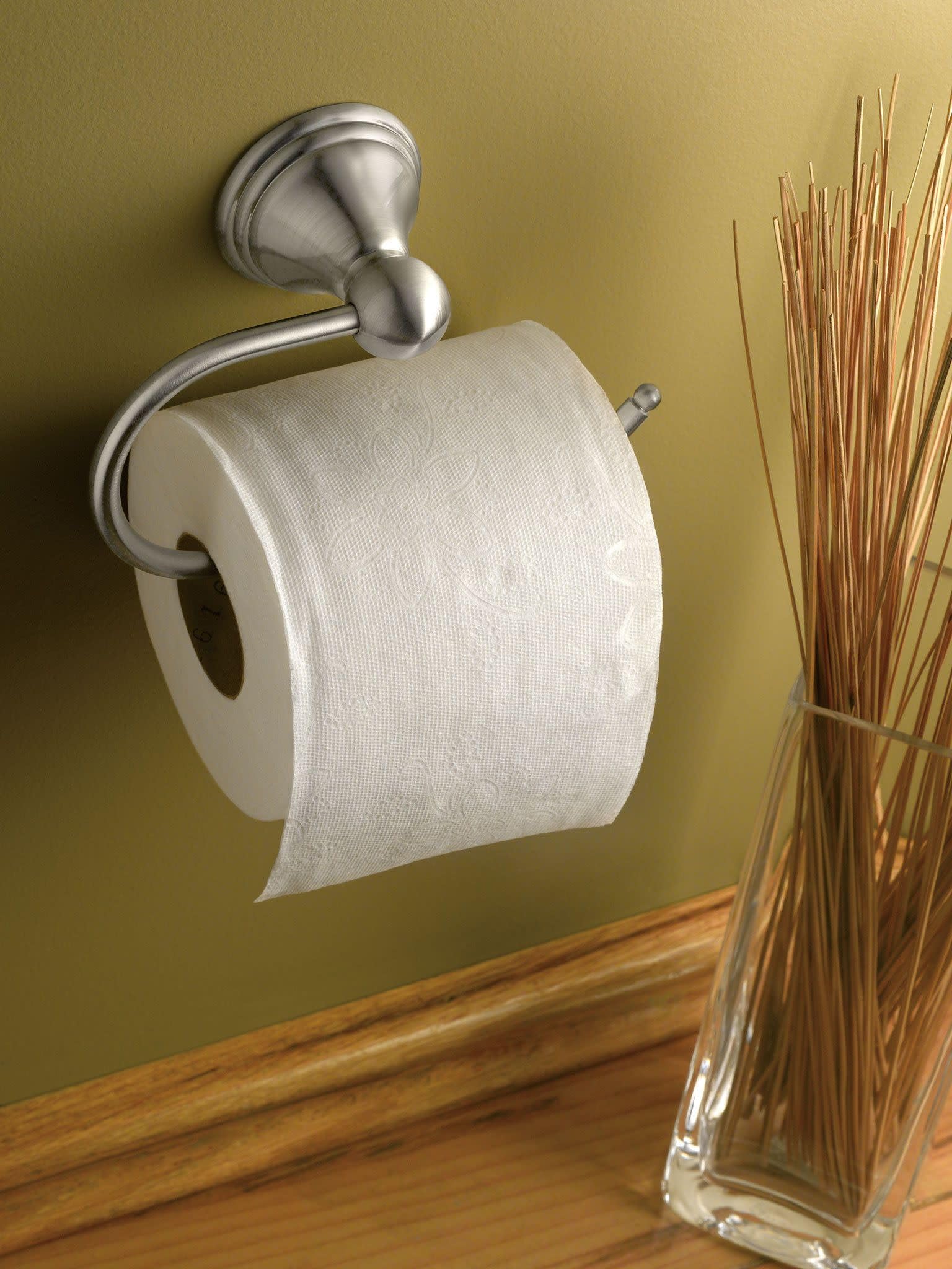Brushed nickel toilet paper holder with curved arm mounted on yellow wall, holding white roll.