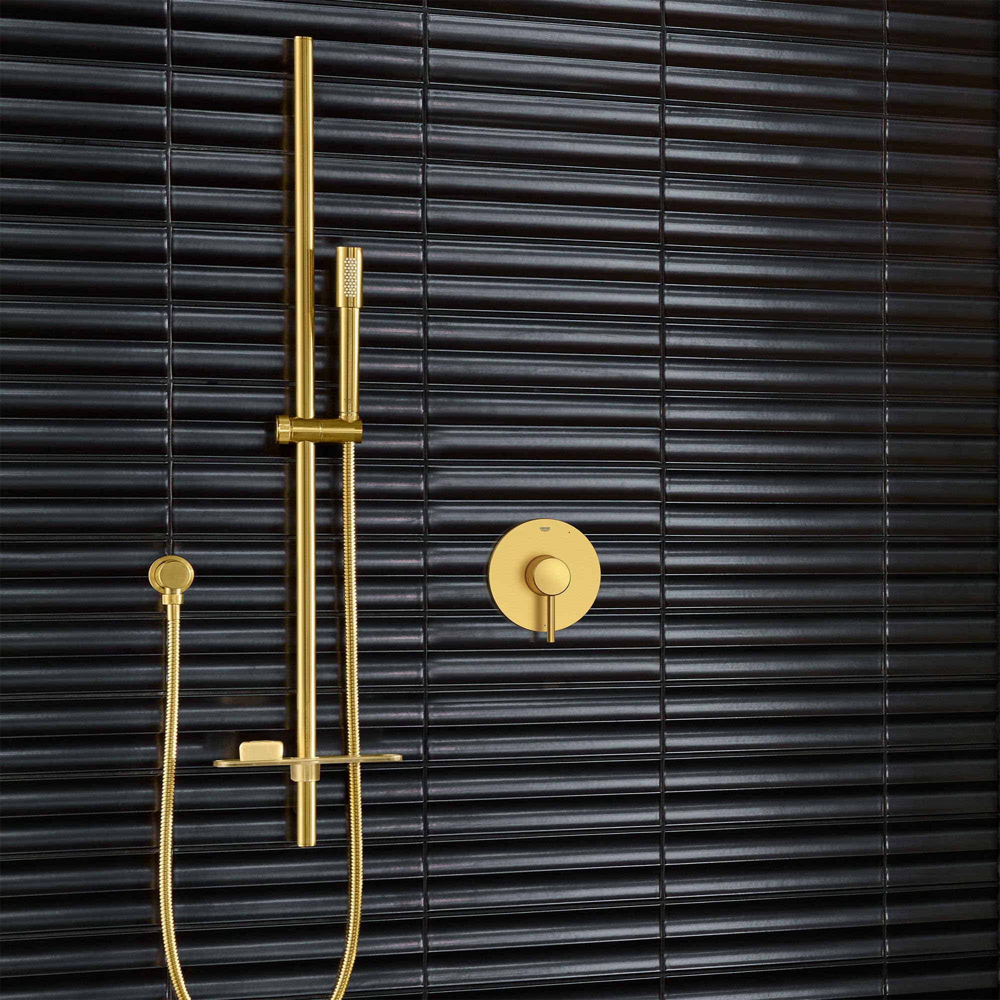 Grohe Concetto brushed gold shower valve trim with hand shower, slider bar, and circular wall outlet mounted on black tile.
