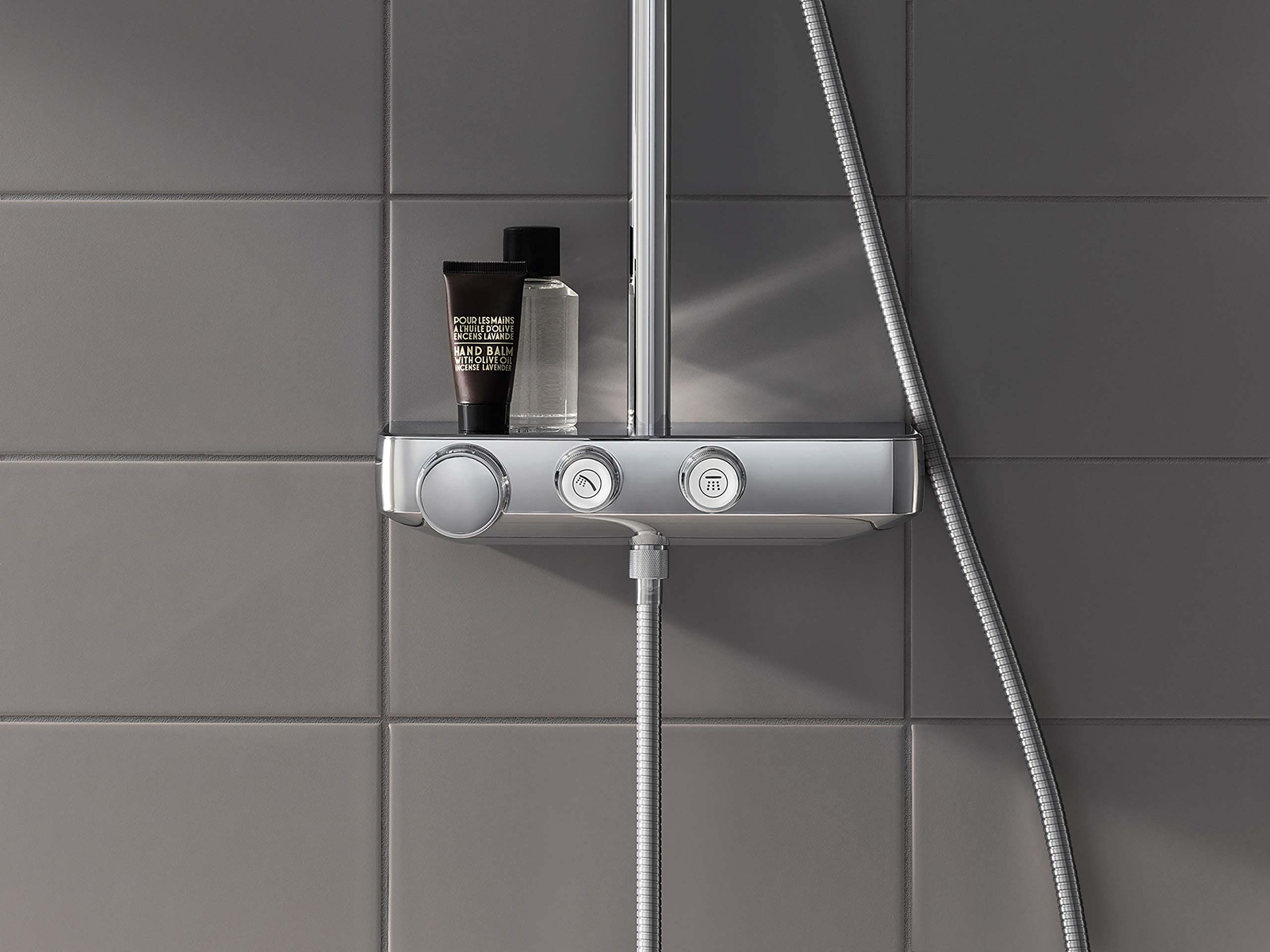 Chrome thermostatic shower system mounted on gray tile wall with control knobs, shelf, and handheld showerhead.
