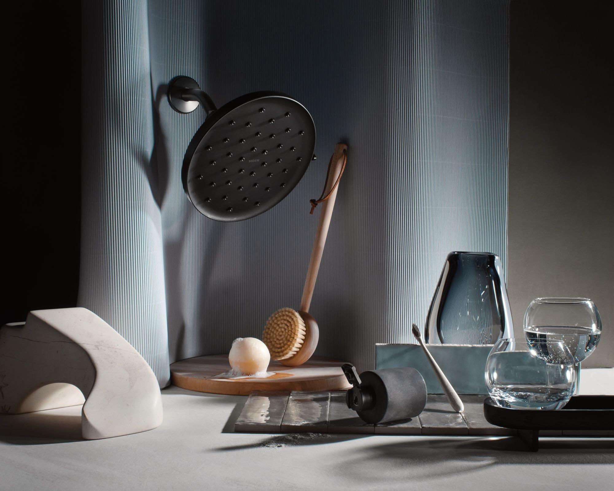 Matte black round rainshower head with multiple spray holes mounted on textured gray wall.