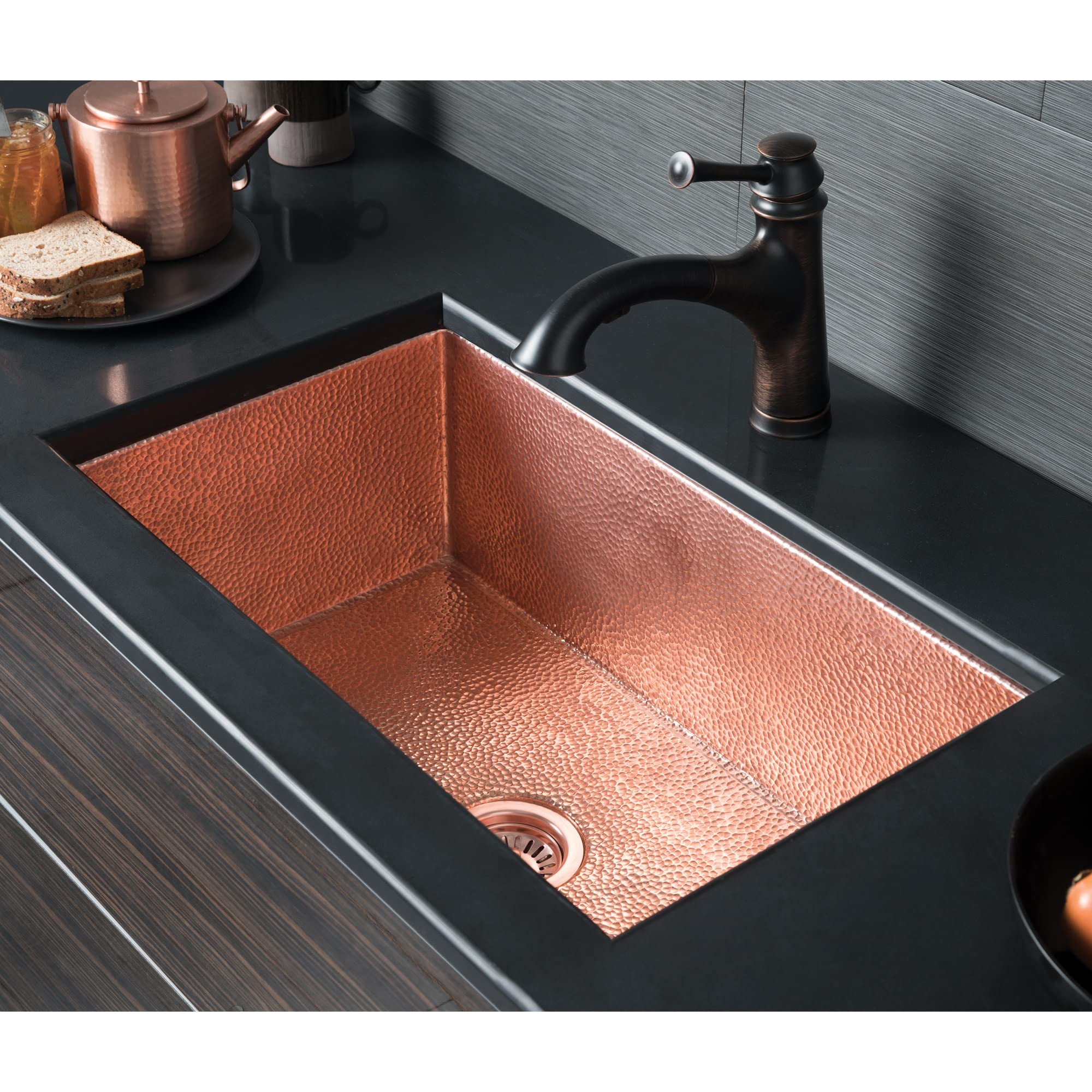 Rectangular copper kitchen sink with hammered texture interior, brushed nickel faucet, and drain visible in dark countertop.