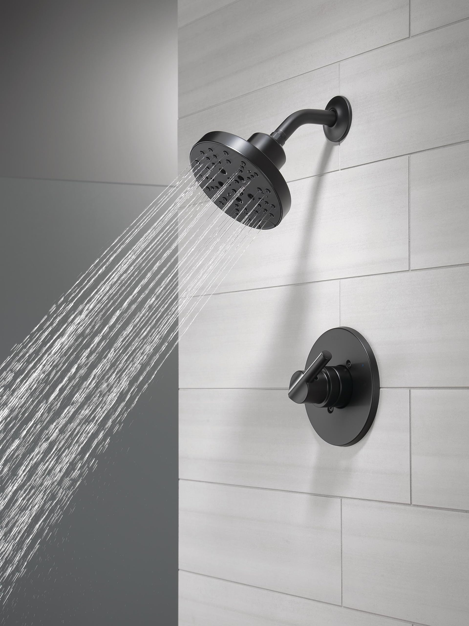 Matte black shower head with curved arm and control valve mounted on white tile wall with water flowing.